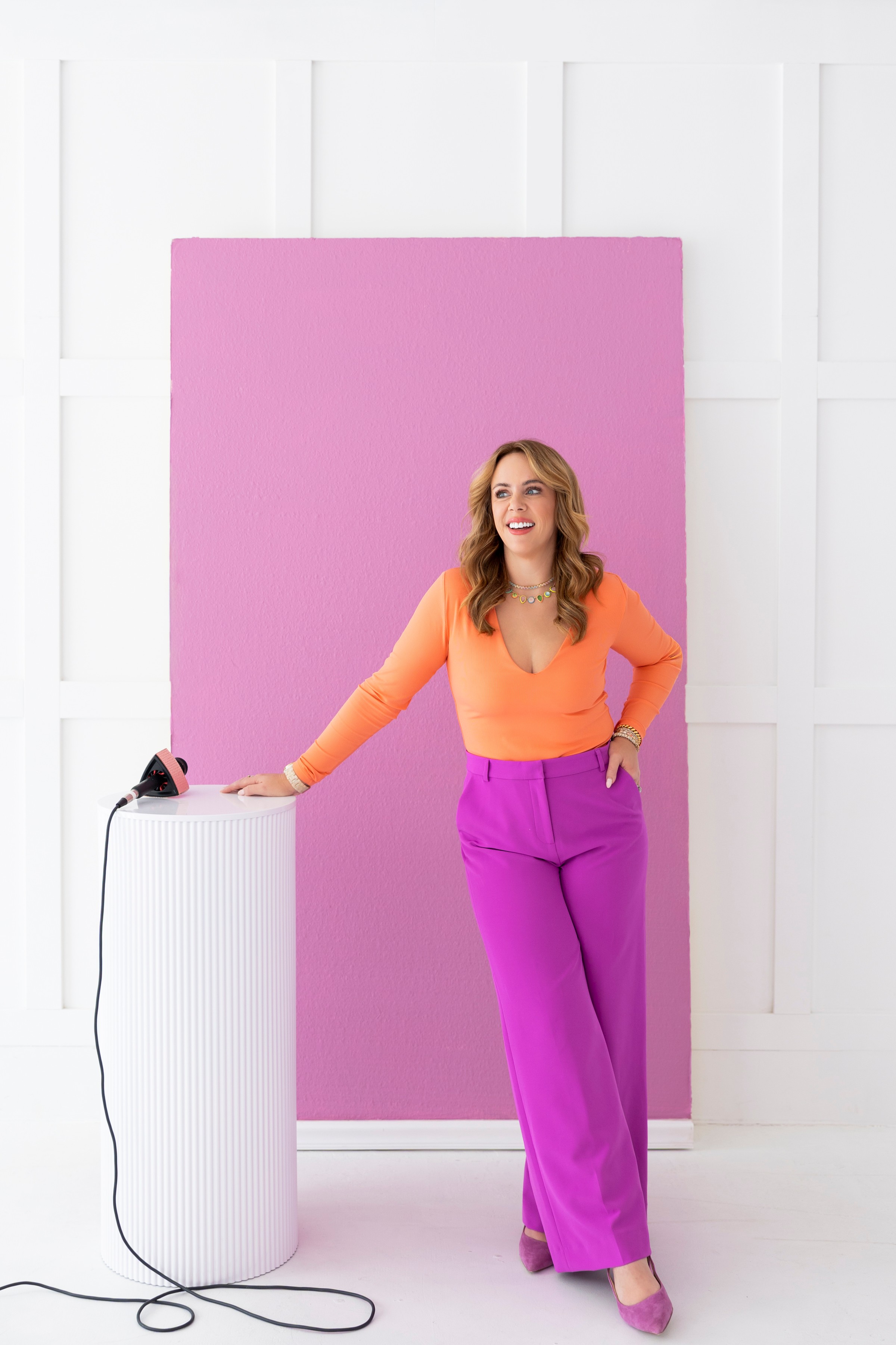 Woman with long hair in orange top and purple pants standing next to a white pedestal with a cord, against a pink and white background.