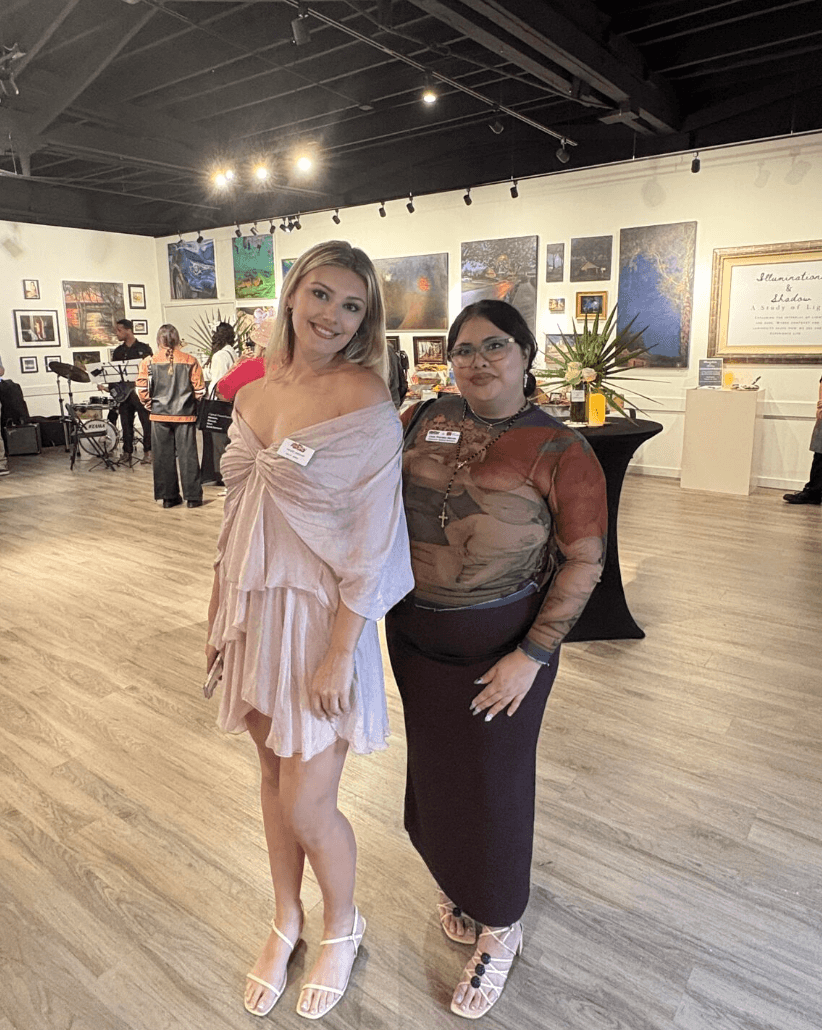 Two women standing in an art gallery, smiling, with artwork displayed on the walls behind them.