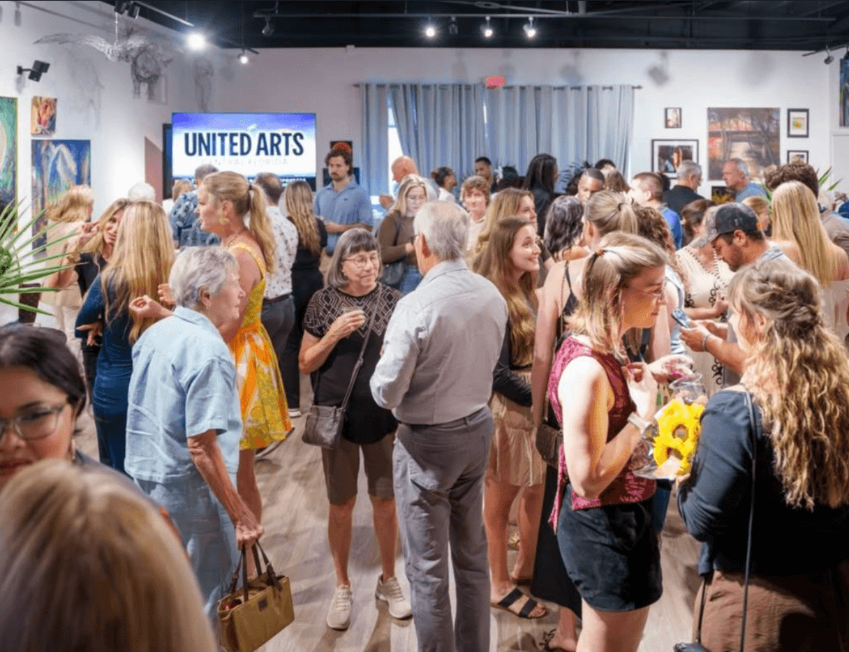 Crowd of people at an art gallery event, with artworks on walls and a sign reading 'United Arts'.