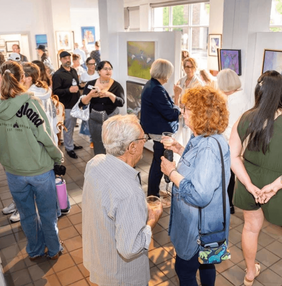 People gather in an art gallery, viewing paintings and talking, with windows and artwork visible in the background.