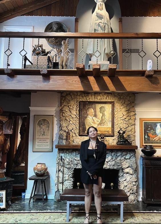 Woman in black blazer and skirt standing in front of a stone fireplace with artwork and decorative items, inside a room.