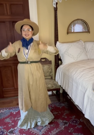 Person dressed as a scout in a room with a bed, wooden furniture, and a red rug, smiling and wearing a hat.