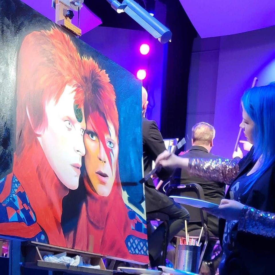 Person with blue hair painting a colorful portrait of two faces on a large canvas in a room with purple lighting.