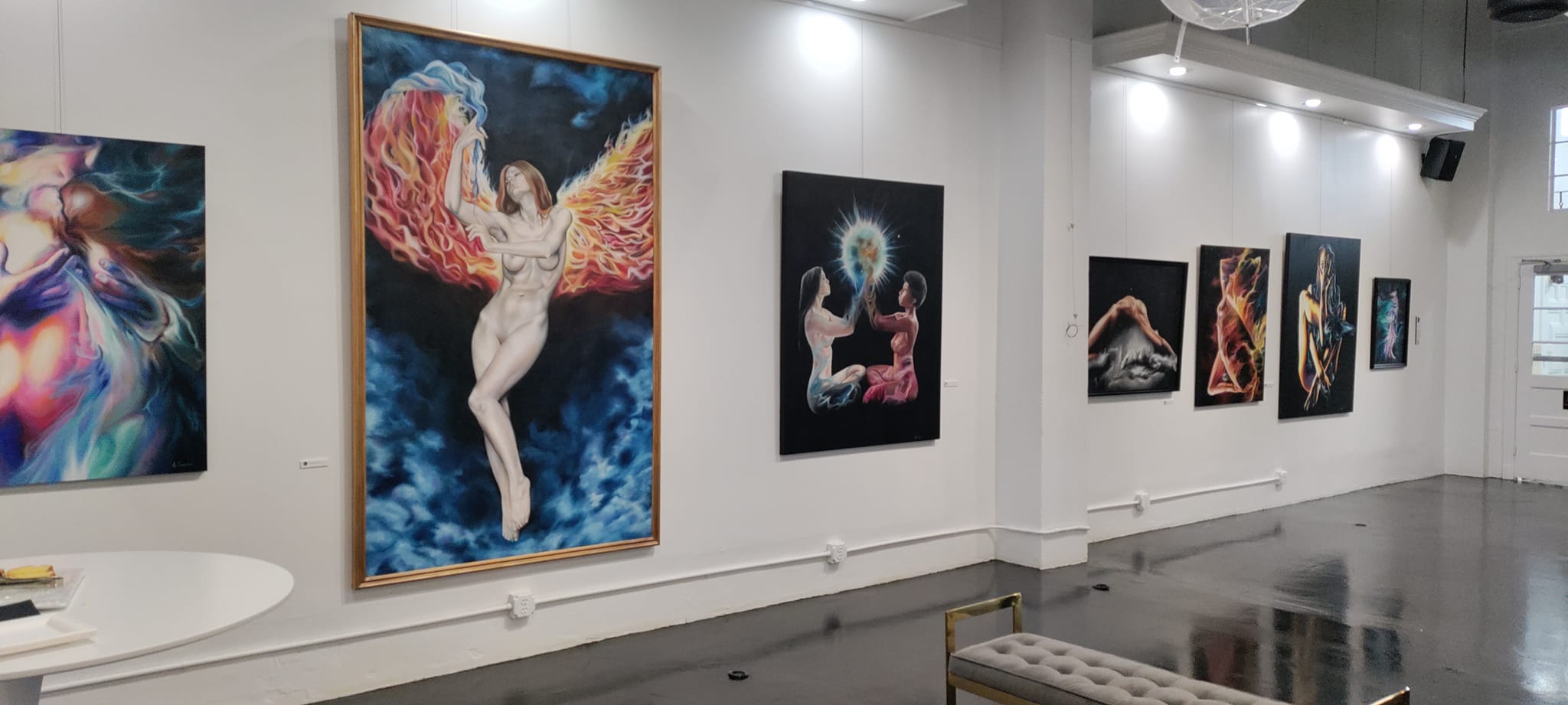 Gallery wall with colorful paintings, including a central artwork of a figure with wings, in a bright, modern space.