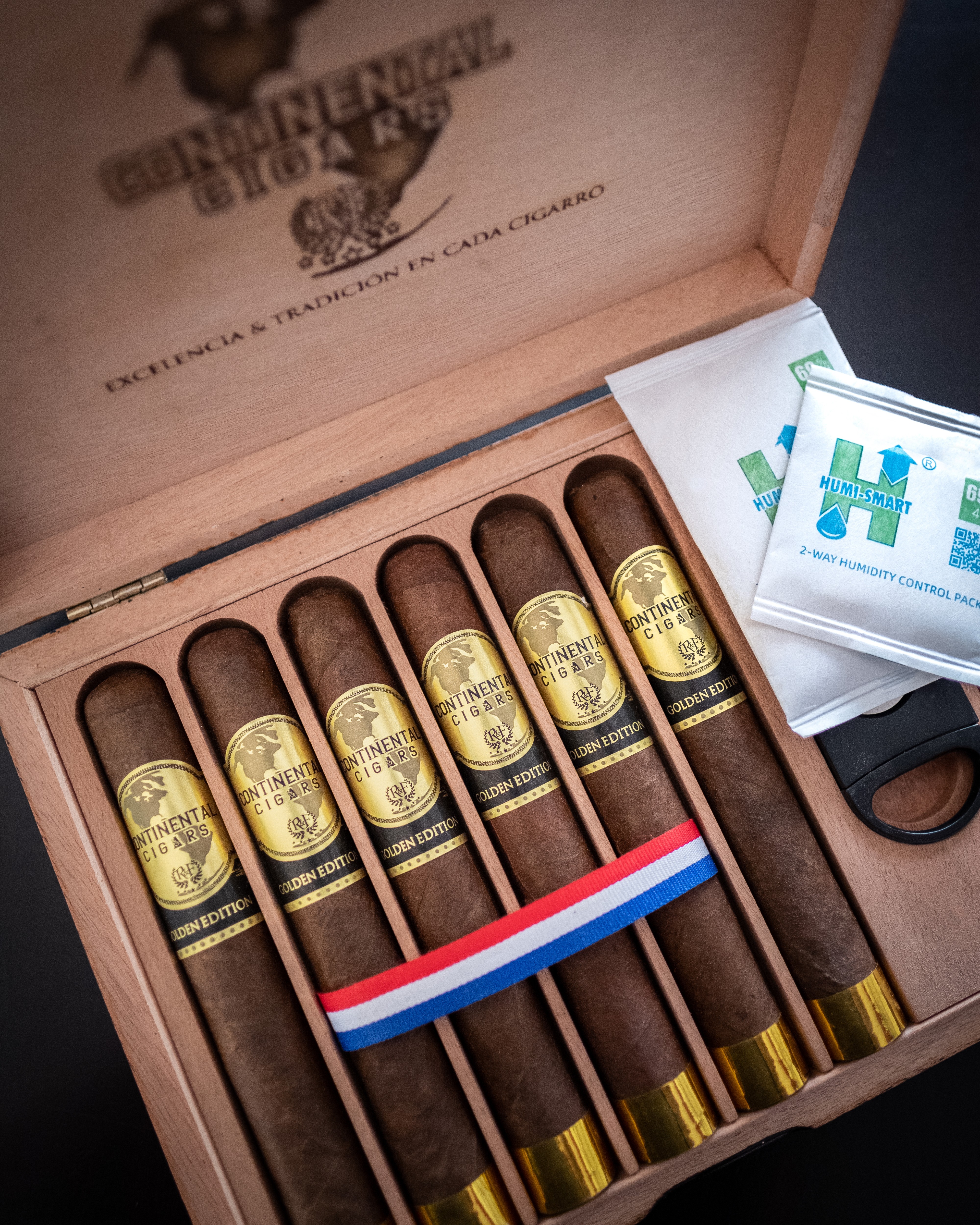 Open wooden box containing seven cigars with gold labels, a ribbon, and a pack of matches inside.