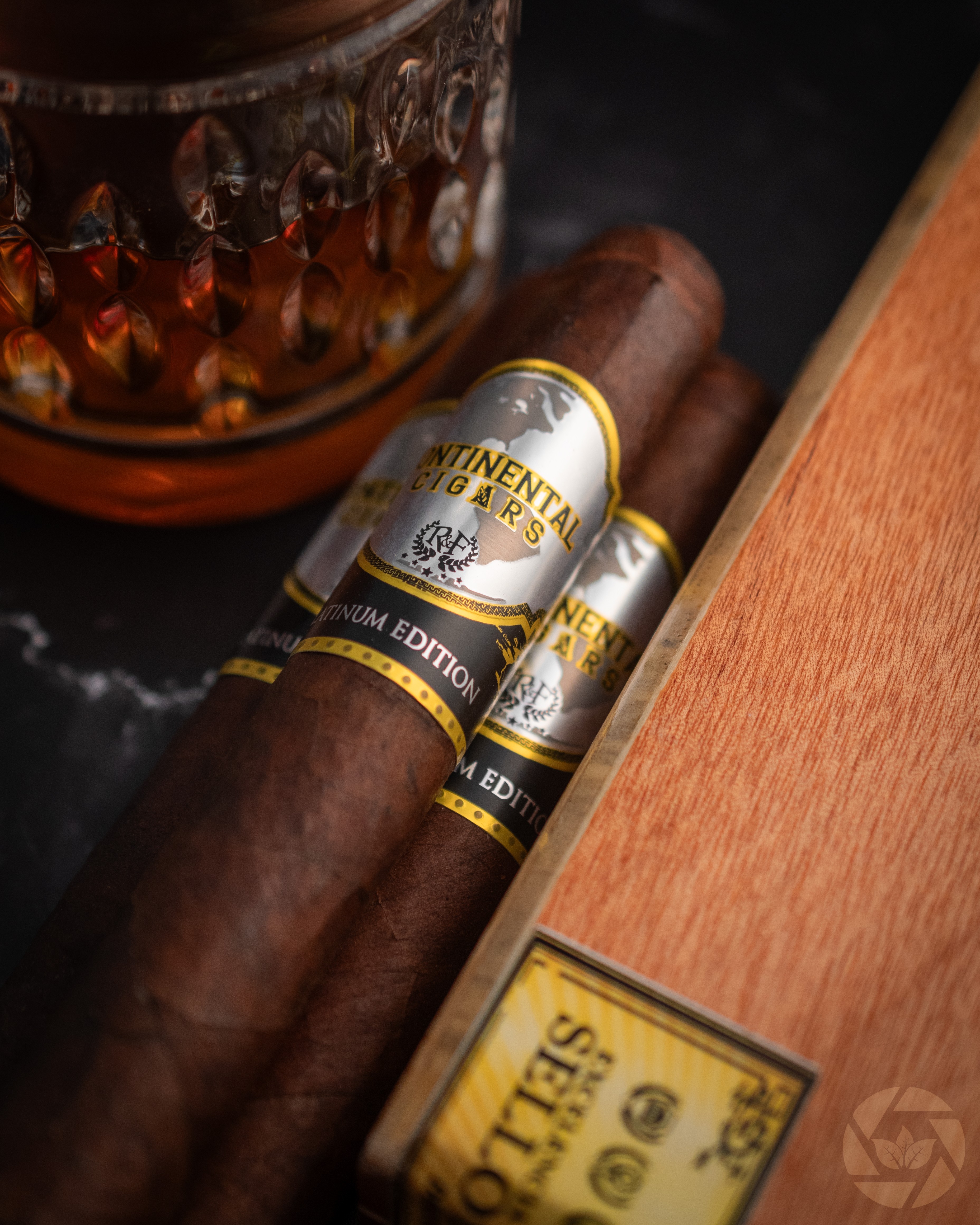 Two cigars with gold and white bands resting on a dark surface next to a wooden box, with a decorative object nearby.