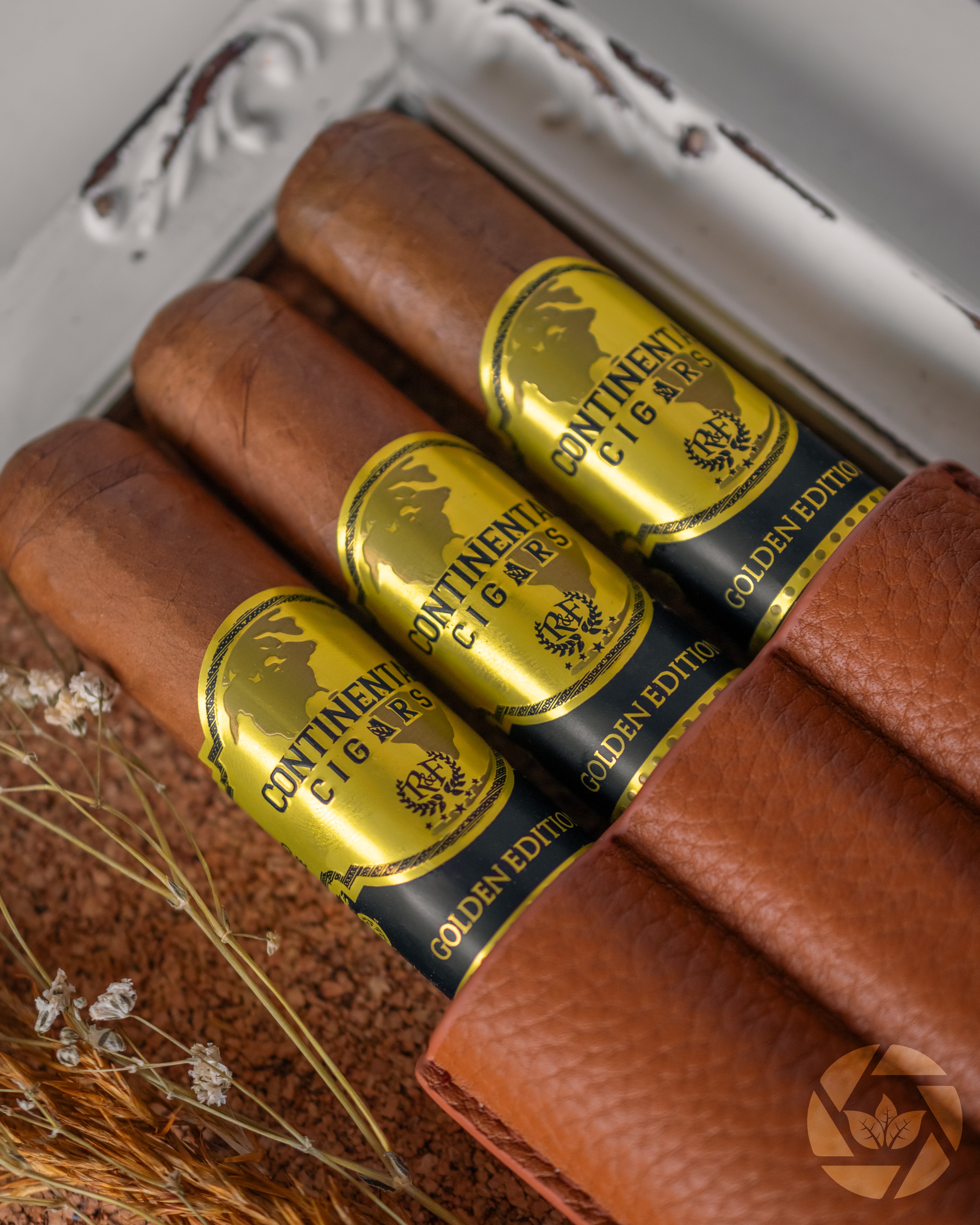 Three cigars with gold and black bands inside a white box, with dried plants nearby.