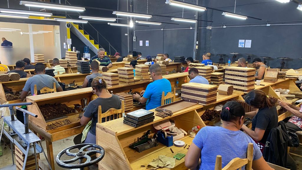 People working at wooden tables in a large, well-lit workshop or factory setting with stacks of materials and tools.