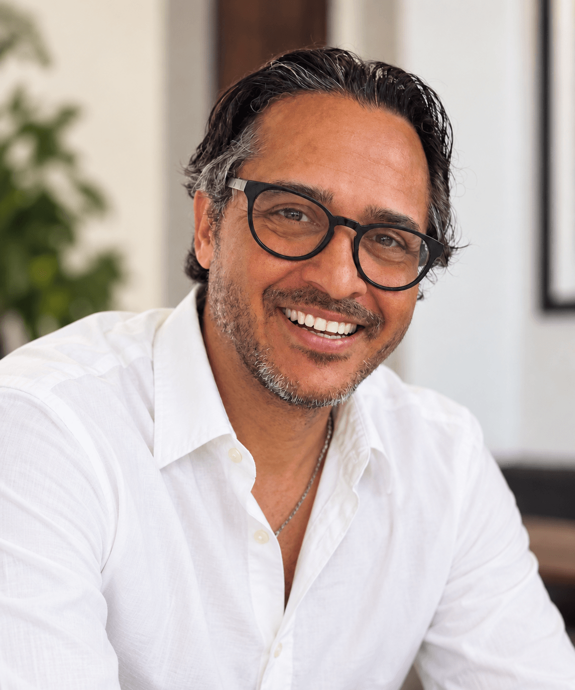 Man with glasses and dark hair, smiling, wearing a white shirt, indoors with a blurred background.