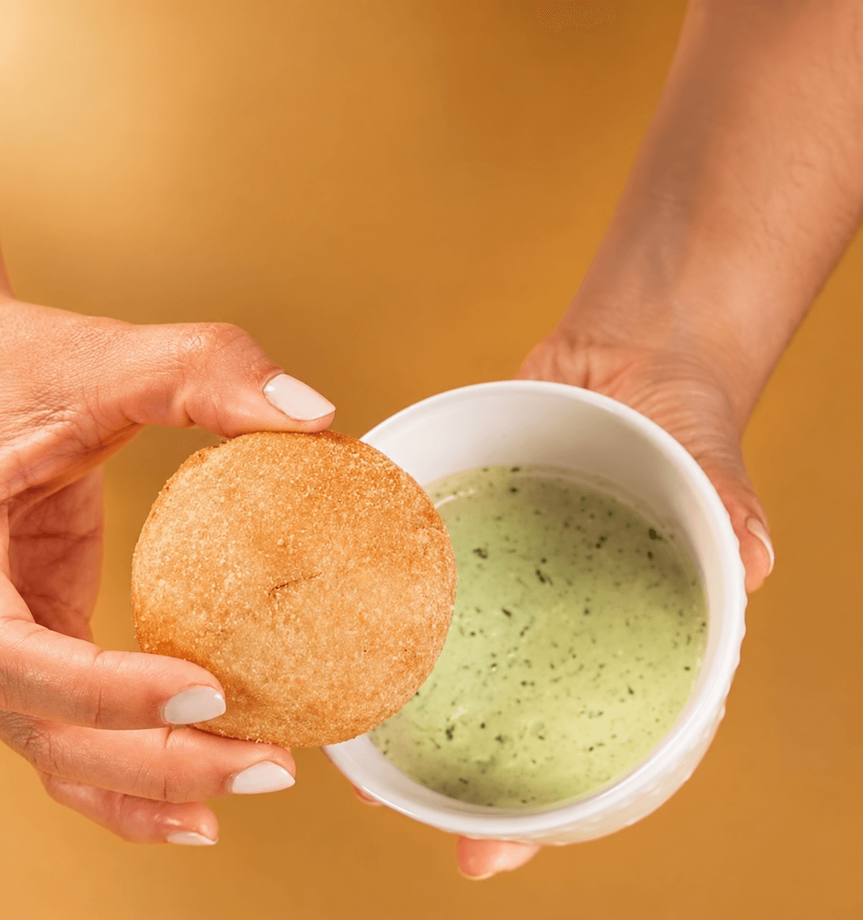 Person holds a round fried snack near a cup of green soup or sauce, with a blurred warm background.