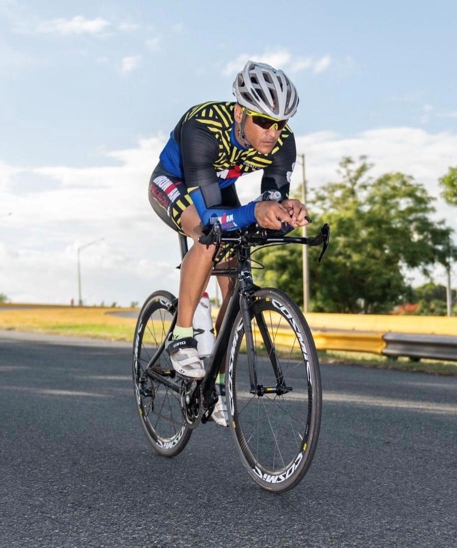Cyclist wearing a helmet and sunglasses riding a road bike on a paved road with trees and sky in the background.