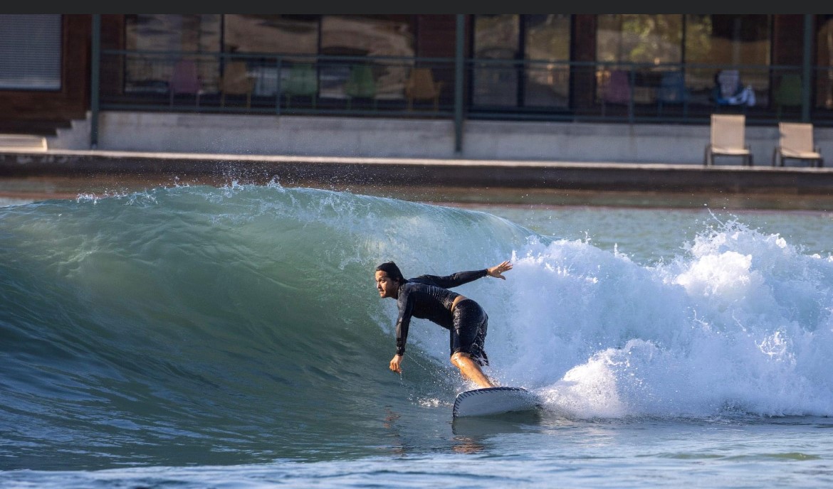 Person surfing on a wave near a building with large windows and outdoor seating area.