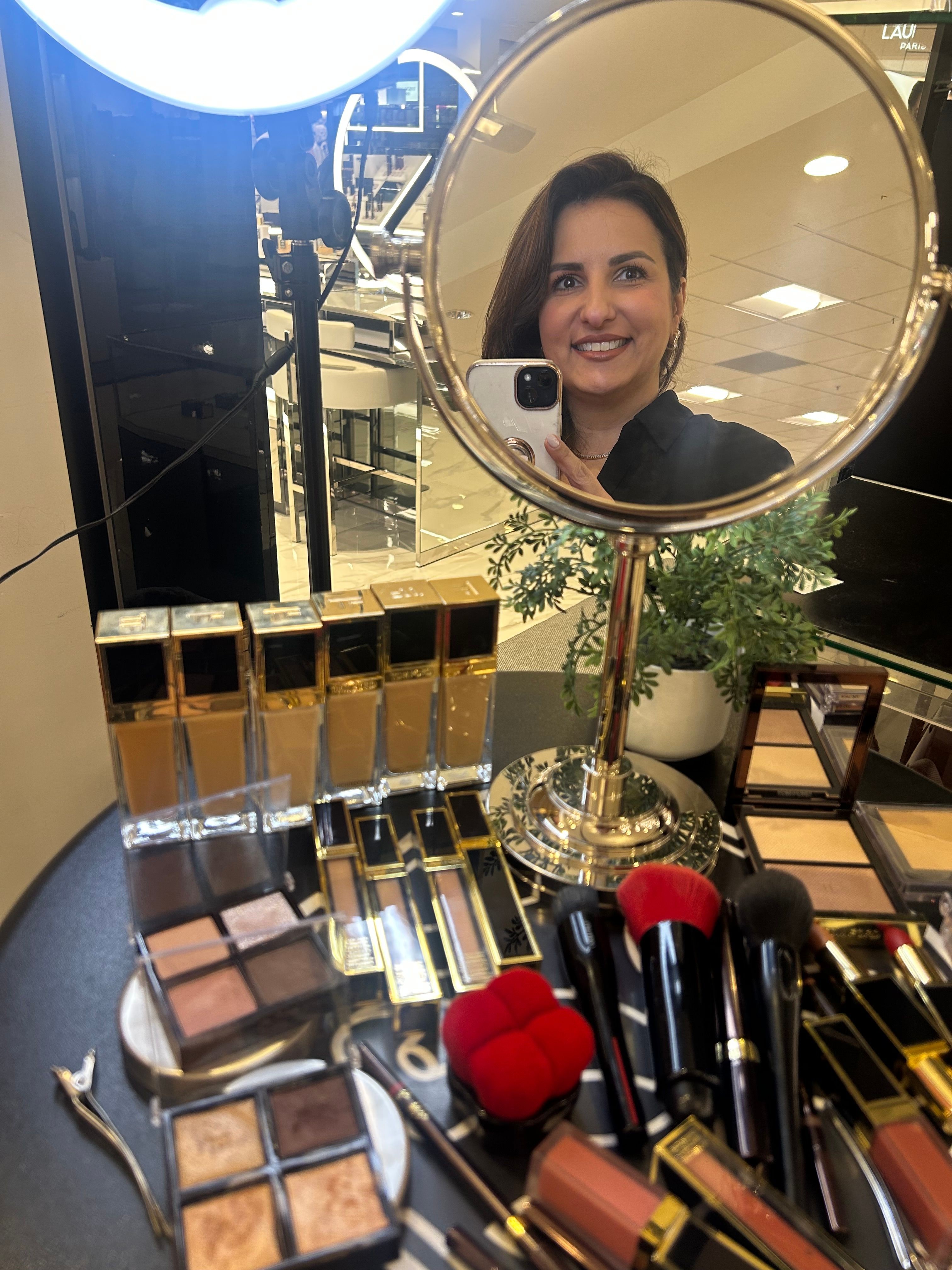 Woman taking a selfie in a mirror, surrounded by makeup products on a table, with a plant and store interior in the background.