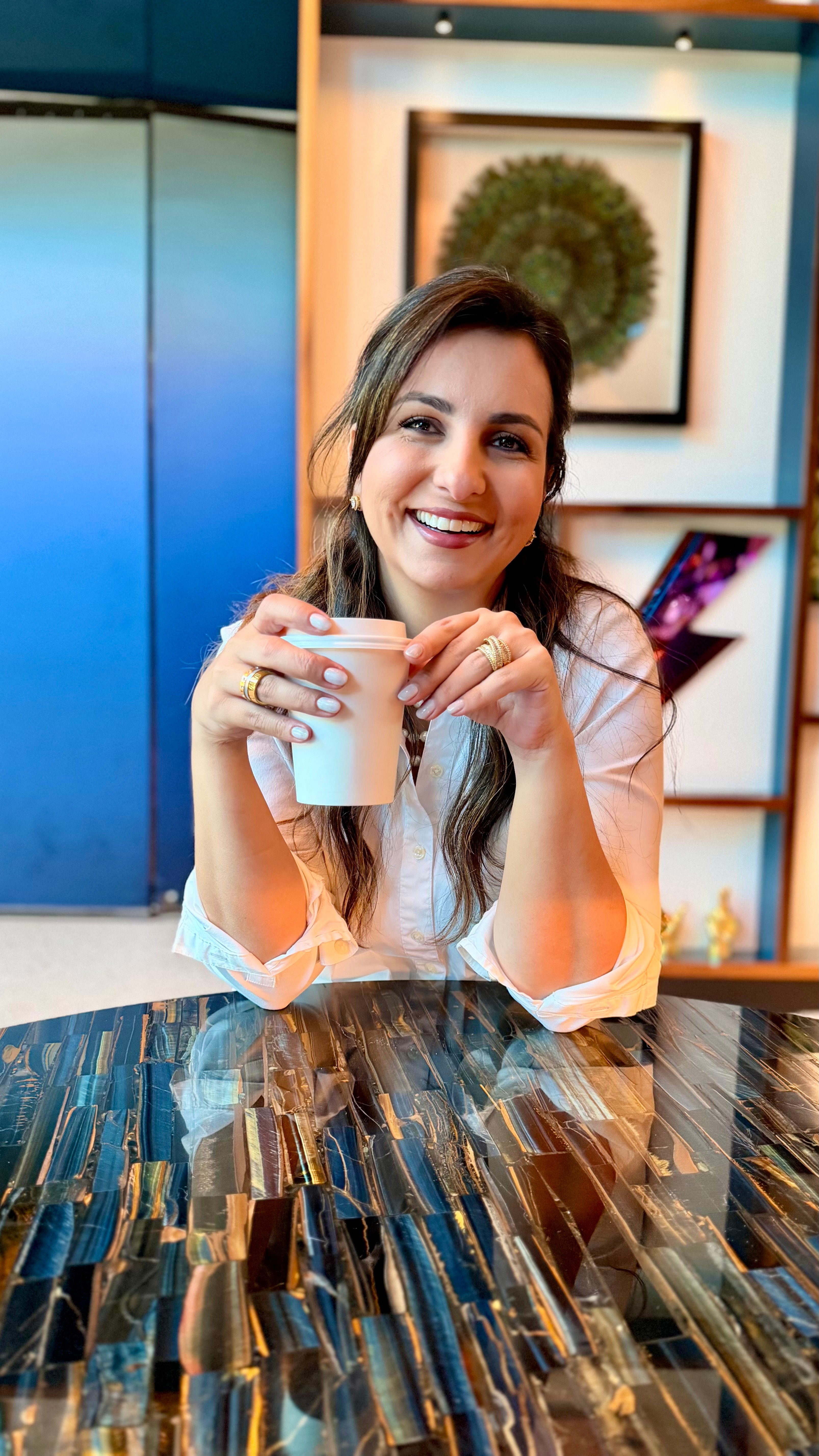 Young woman with long hair smiling and holding a white mug, sitting at a table in a colorful room.