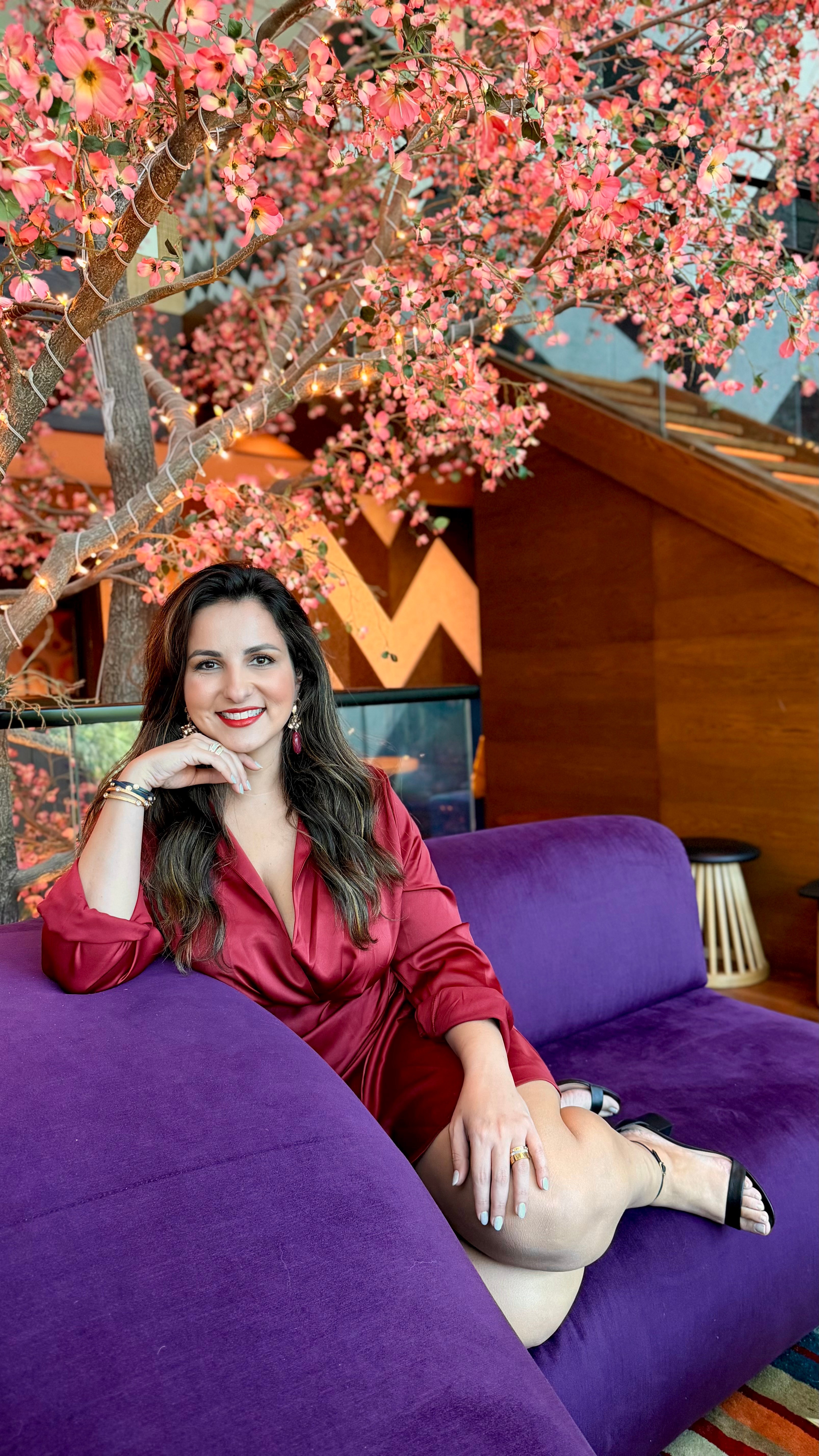 Woman in red dress sitting on purple sofa with pink flowering tree in background.