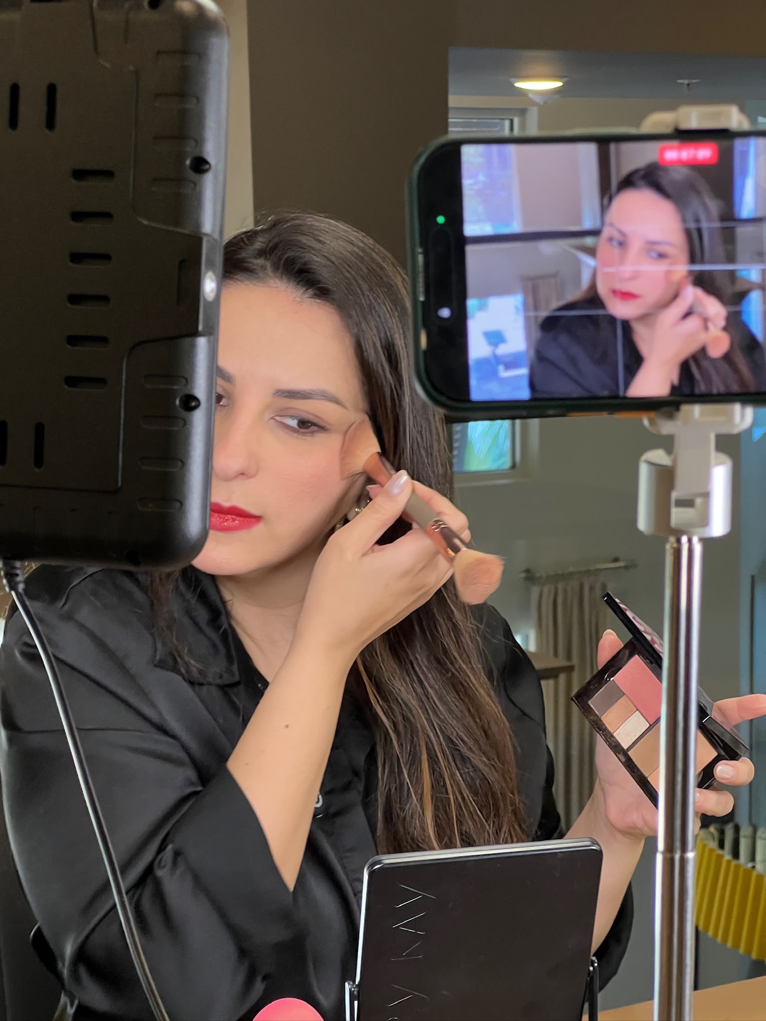 Woman applying makeup while recording herself with a phone, with a camera monitor showing her reflection, in a room.