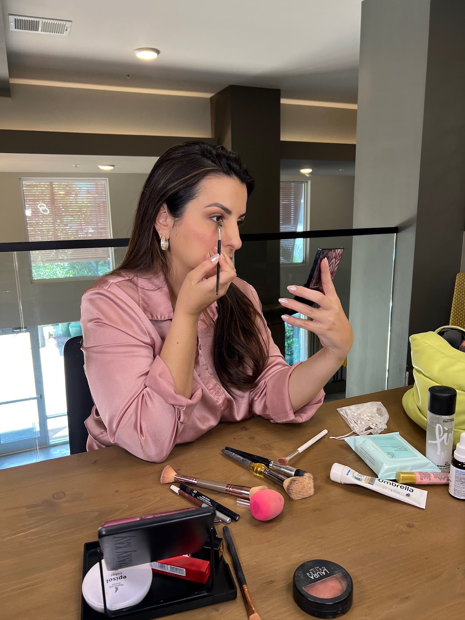 Woman applying makeup at a table with various cosmetics and tools, in a modern indoor setting.