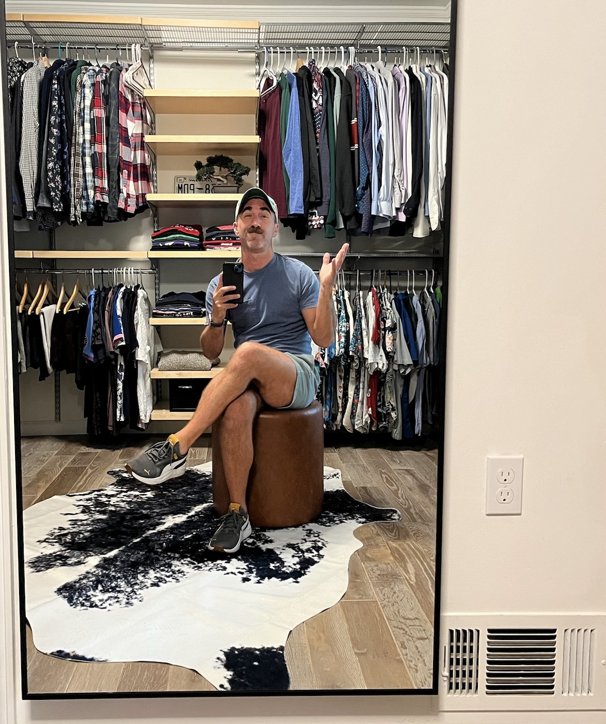 Person sitting on a round ottoman in front of a closet mirror, taking a selfie, with clothes hanging behind, on a cowhide rug.
