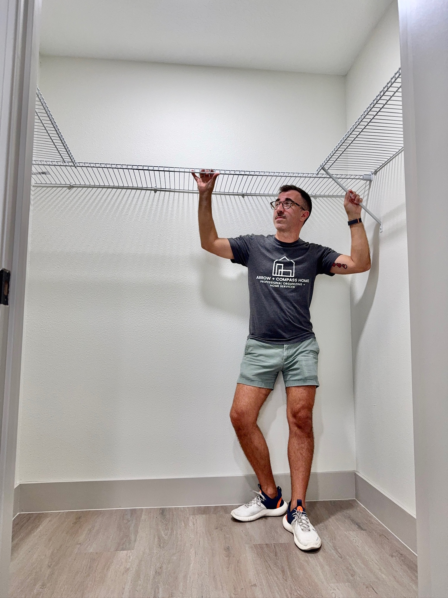 Person standing in a closet with wire shelves, wearing a dark t-shirt, shorts, and sneakers, holding the shelf with both hands.