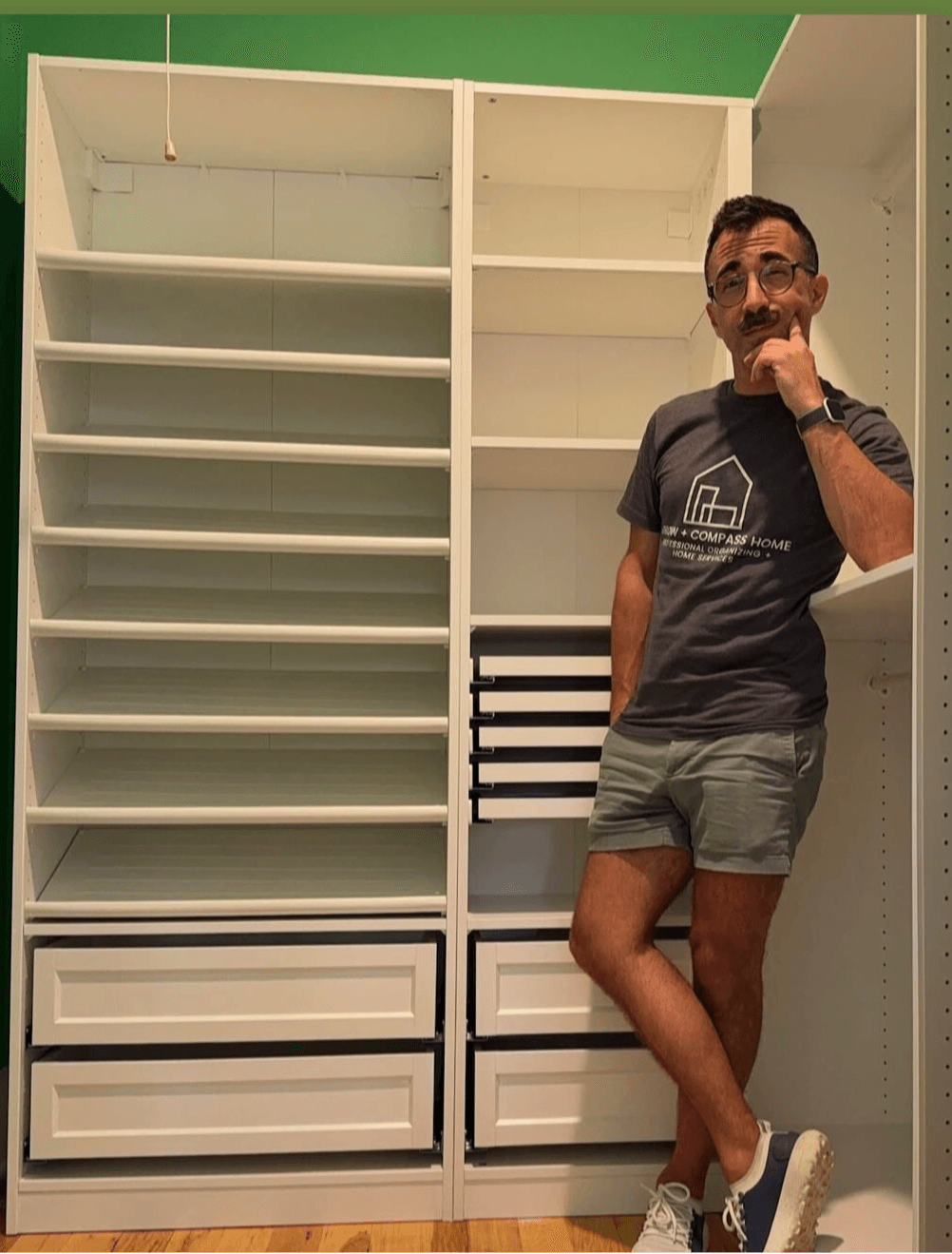 Person standing in front of empty white shelves, wearing a dark t-shirt, shorts, and sneakers, with one hand on chin.