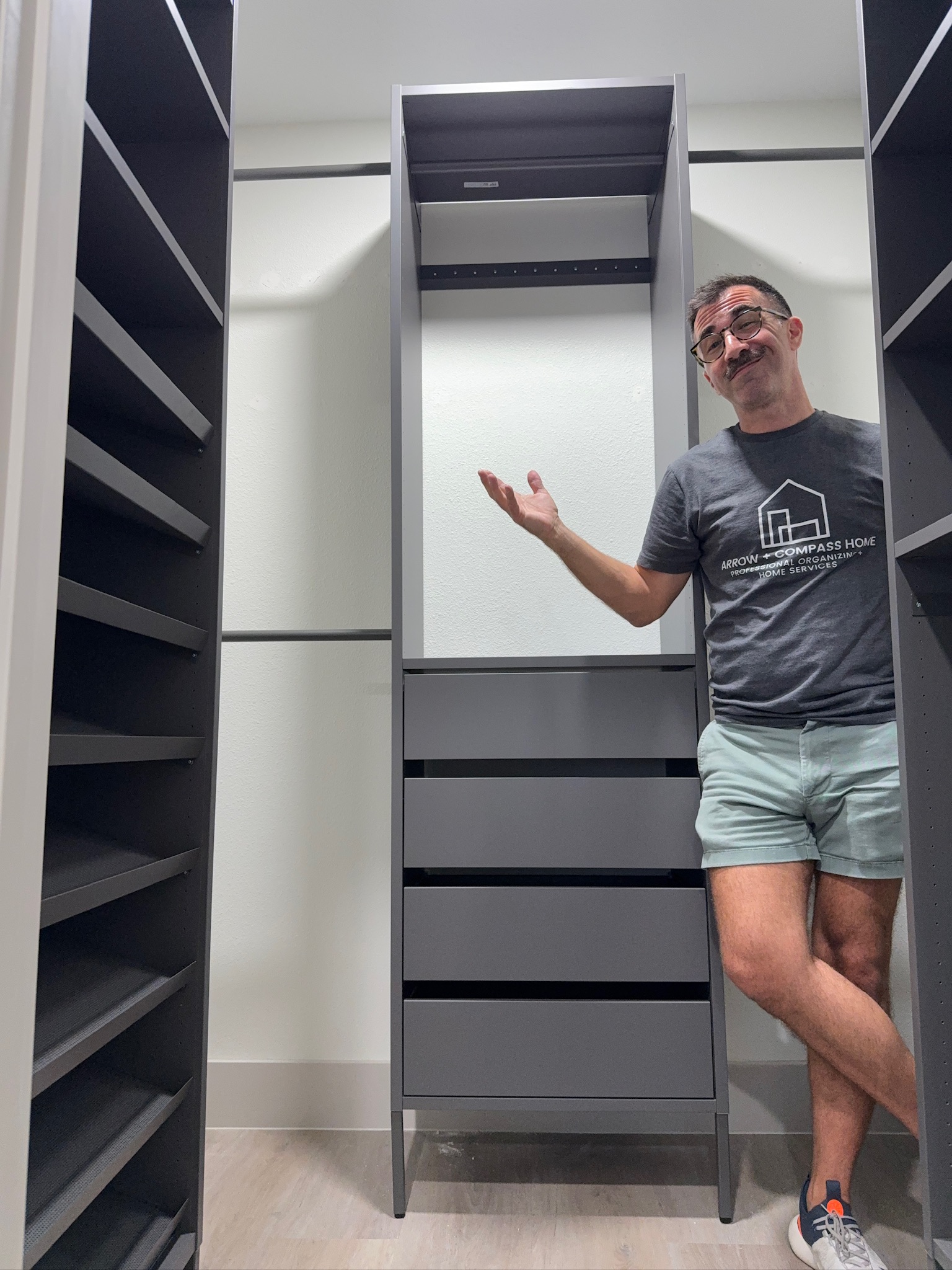 Person standing in a walk-in closet with empty shelves, gesturing with one hand, wearing a gray t-shirt and shorts.