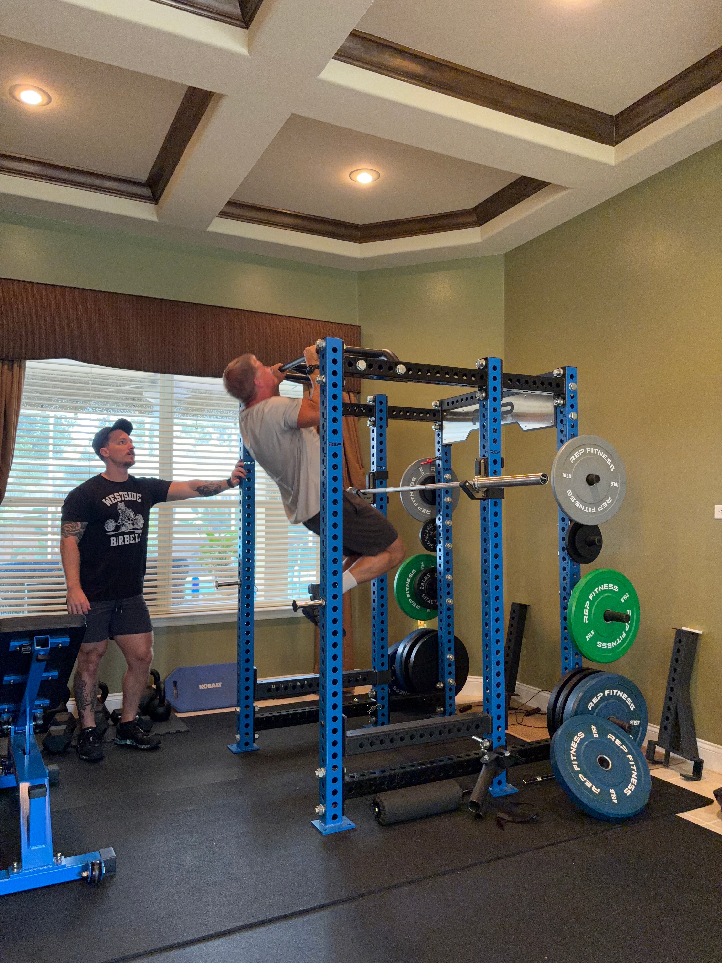 Person performing pull-up on a blue and black gym rack, with a person spotting nearby, in a room with green walls and windows.