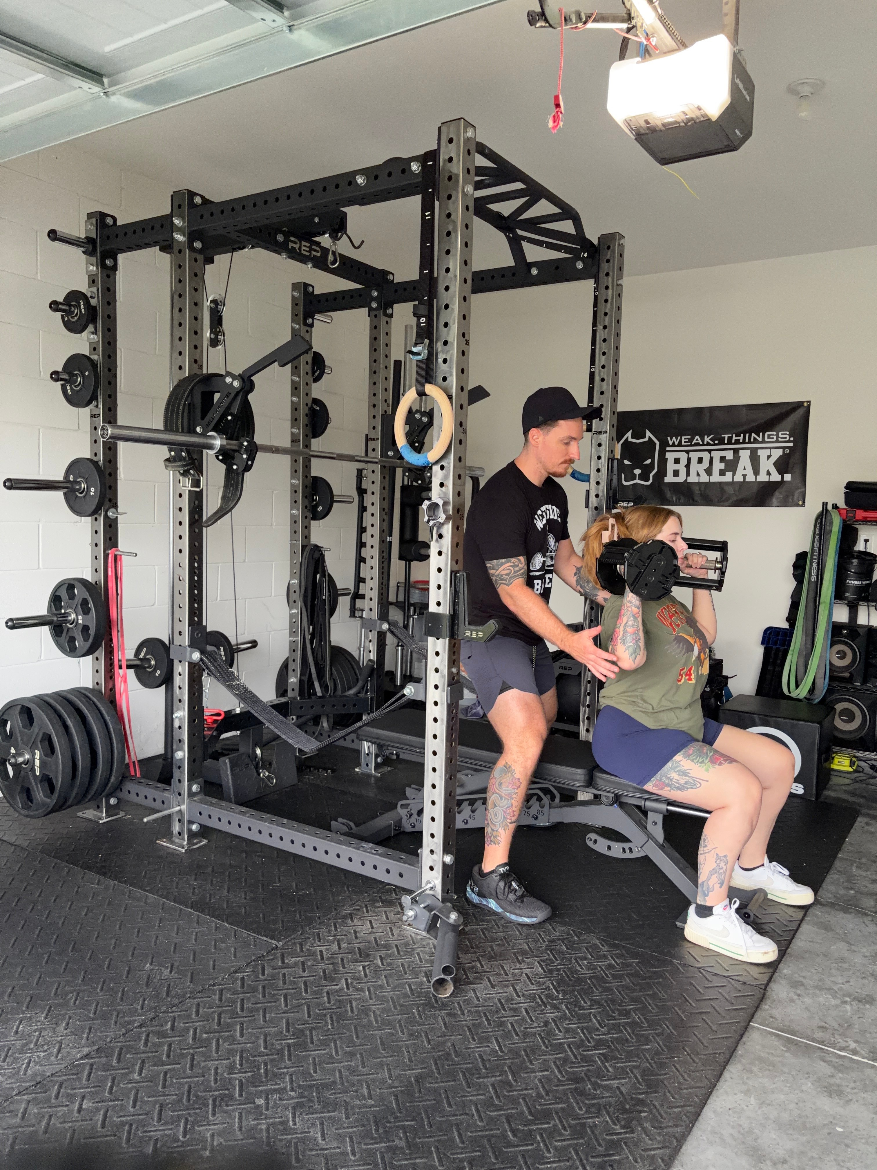 Two people exercising with a barbell in a gym, surrounded by weightlifting equipment and a black mat.