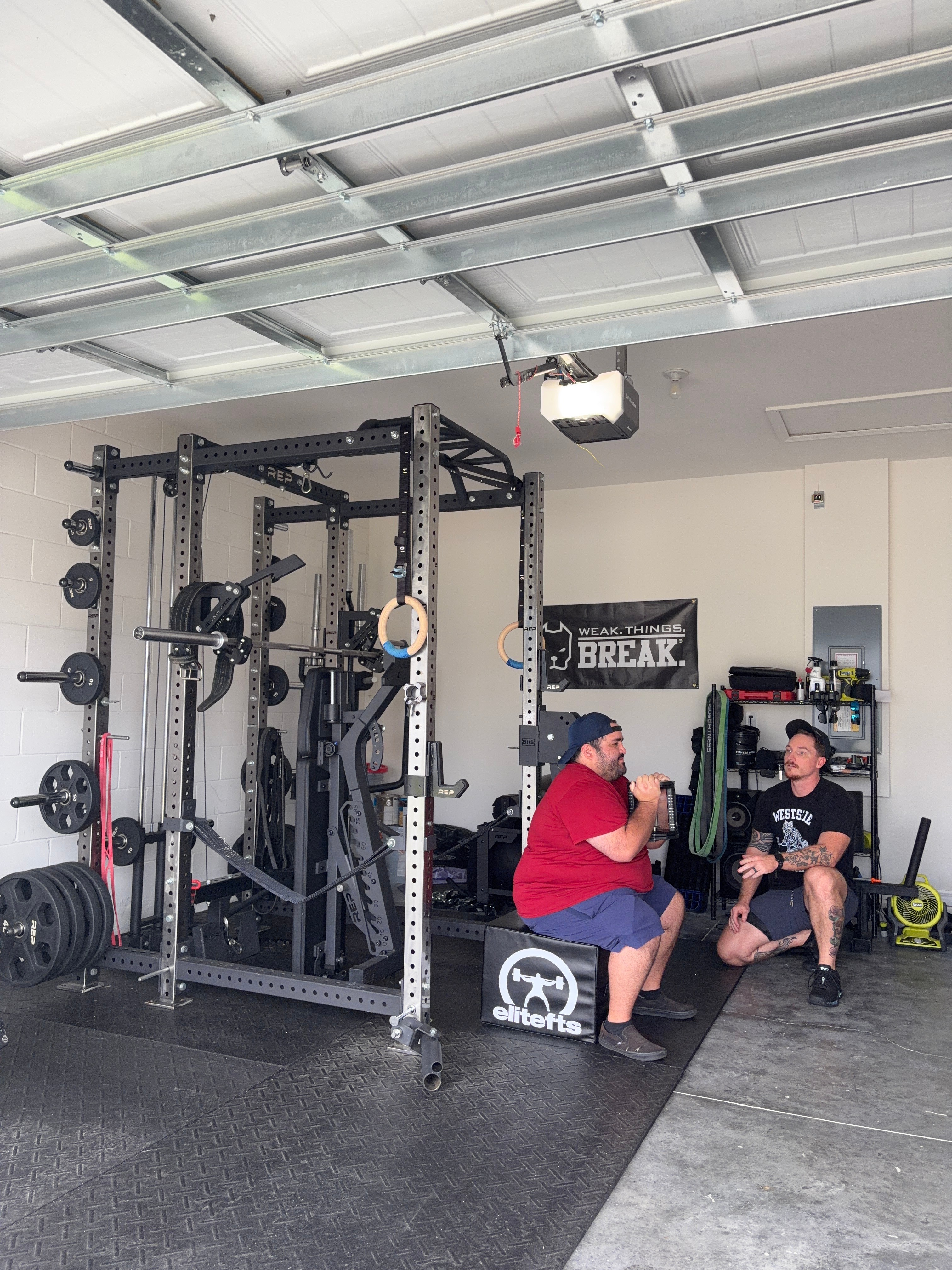 Two people sitting and talking in a gym with weightlifting equipment and a sign that says 'BREAK'.