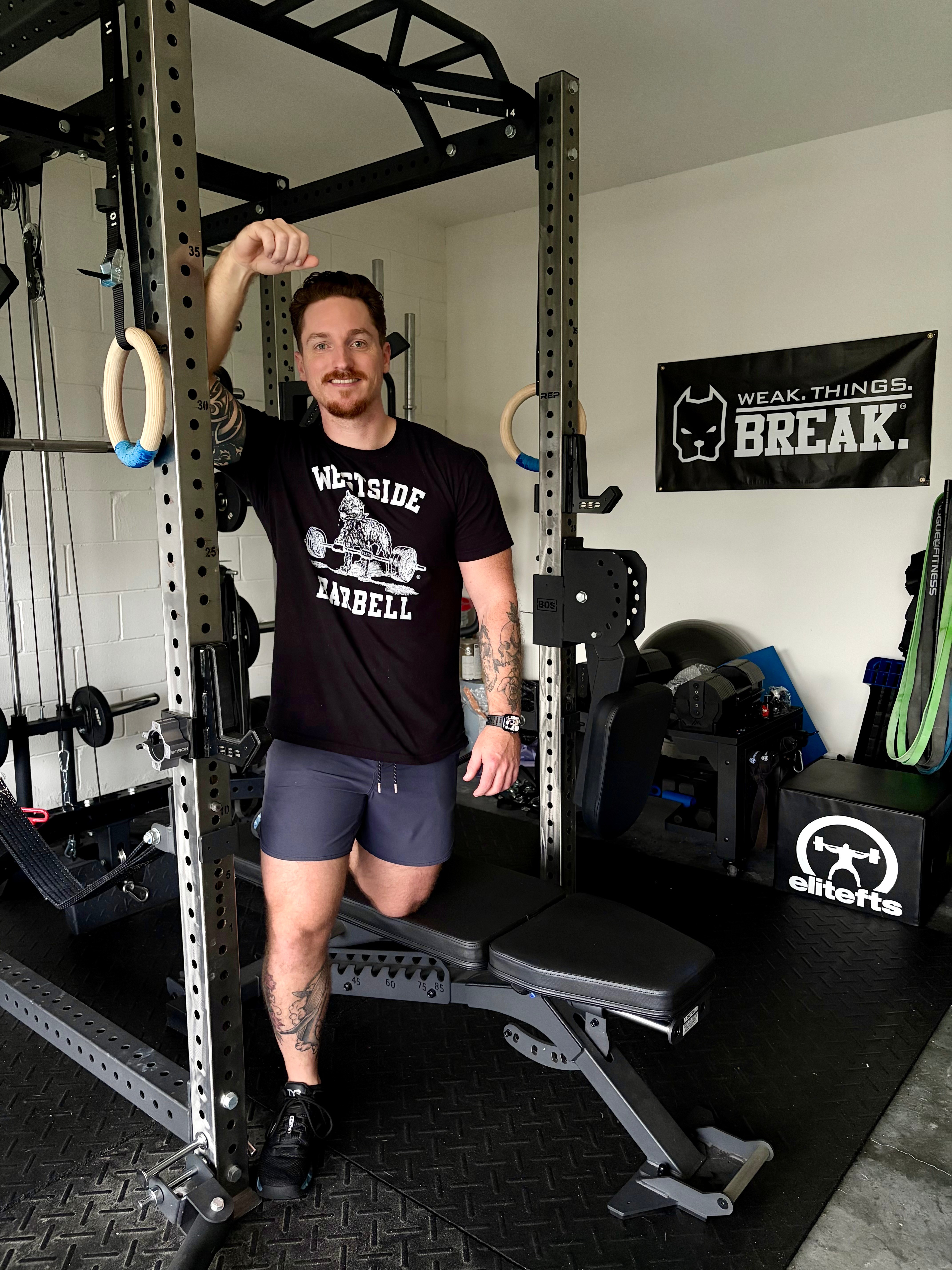 Man standing in gym, smiling, holding gymnastic rings, wearing black t-shirt and shorts, with workout equipment around him.