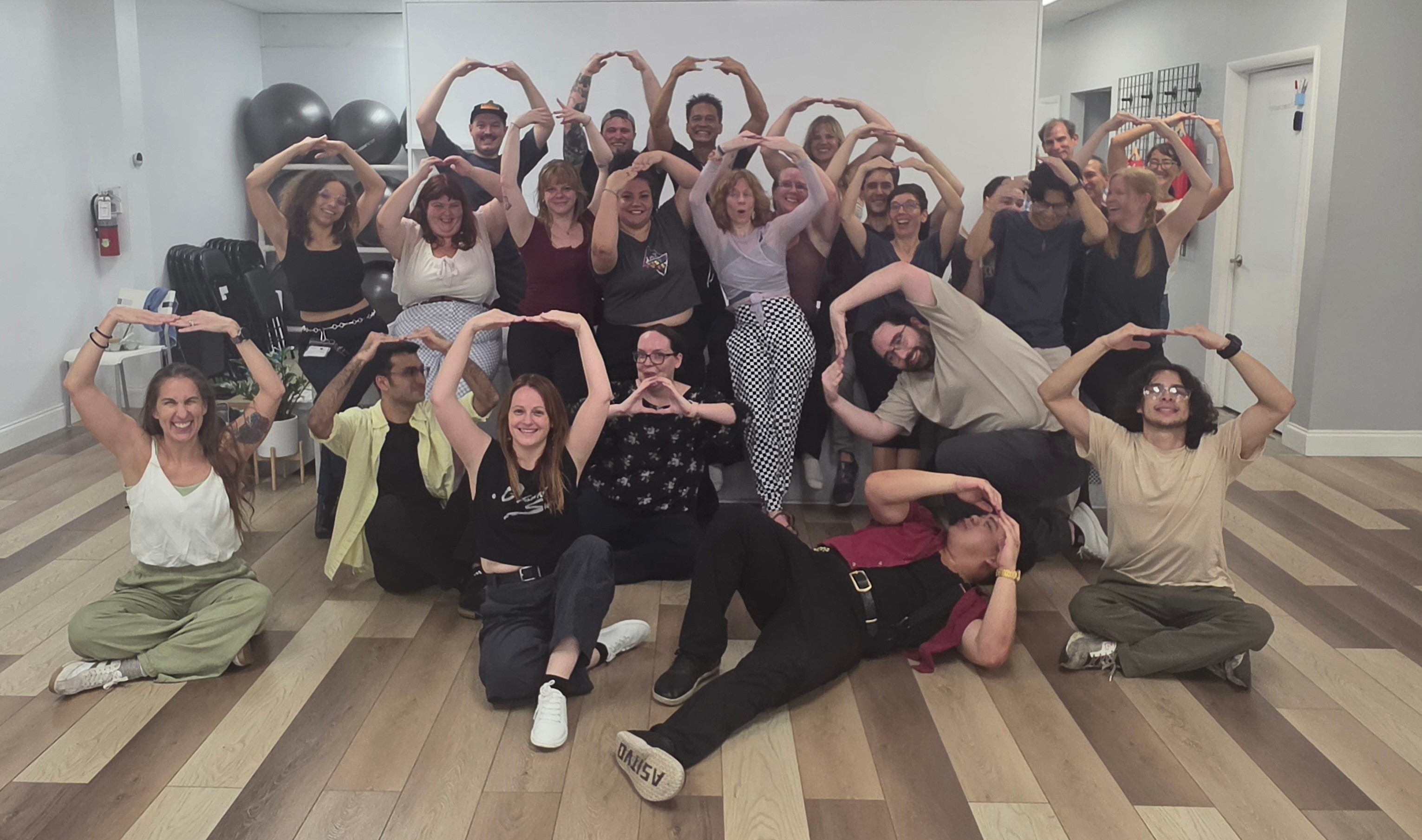 Group of people in a room, some sitting and some standing, forming heart shapes with their arms, smiling.