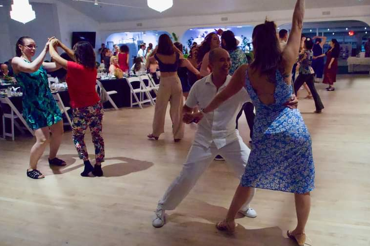 People dancing in a spacious room with a wooden floor, some seated at tables in the background.