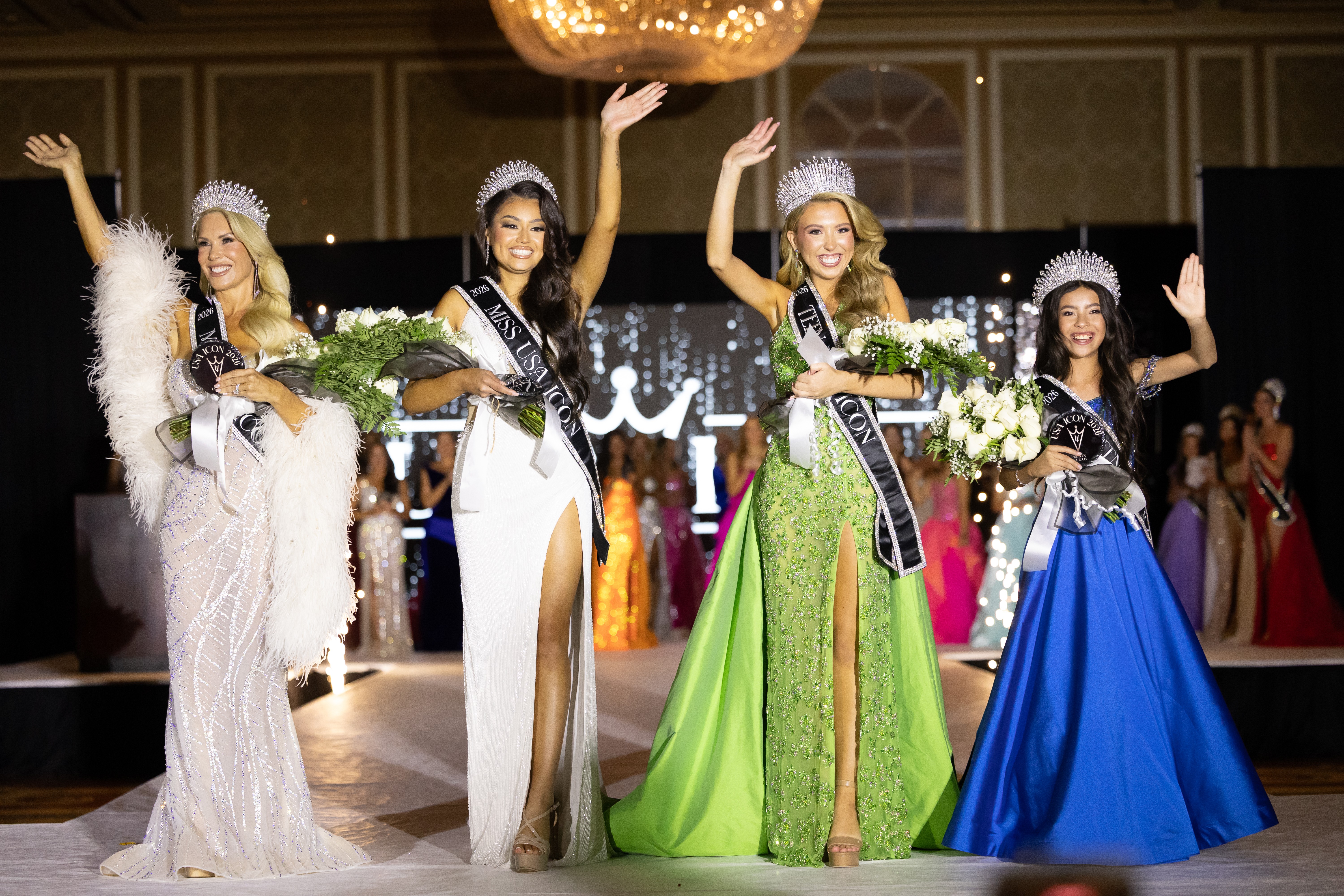 Four women in evening gowns and crowns on stage, holding bouquets, waving, with a decorated background and other contestants in the background.
