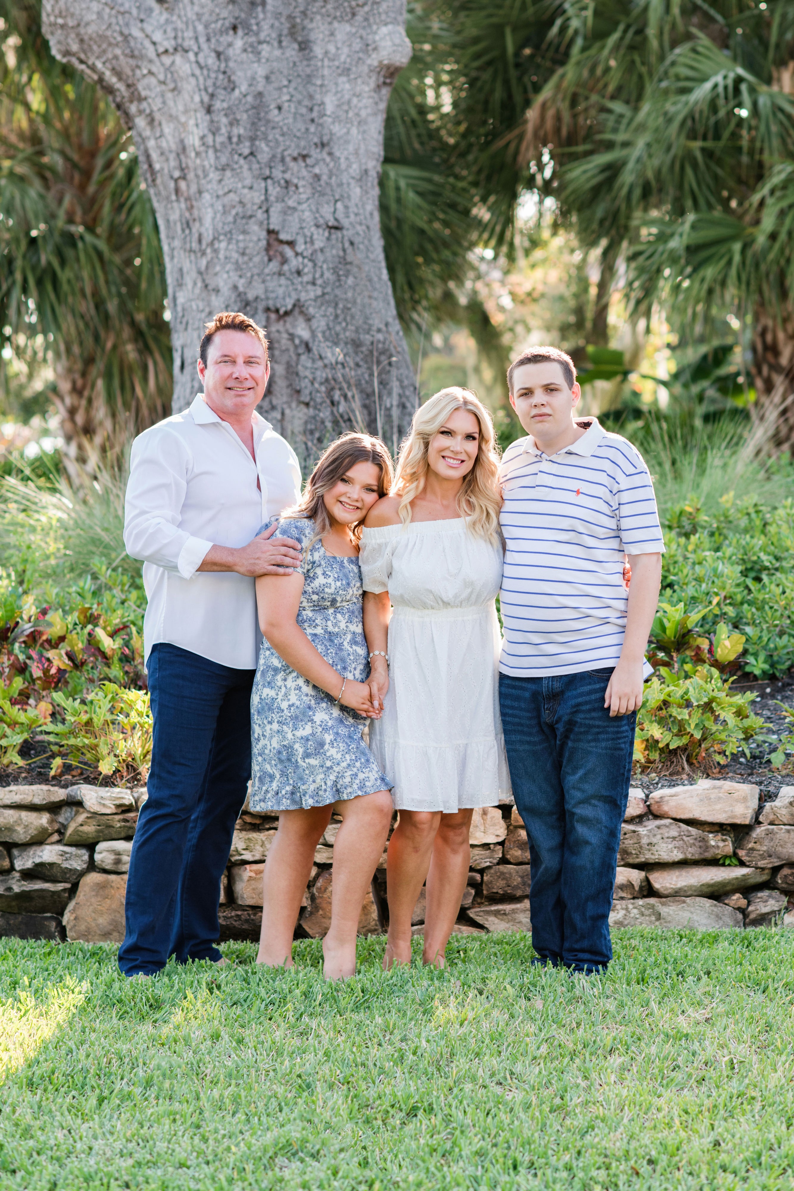 Four people standing outdoors in front of a large tree and greenery, smiling, with a stone border behind them.