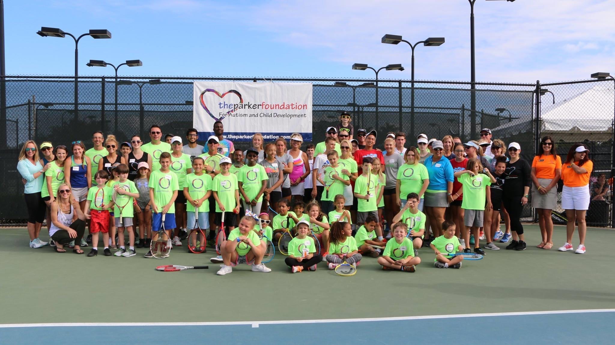 Group of children and adults on a tennis court, some wearing bright green shirts, with a banner in the background.