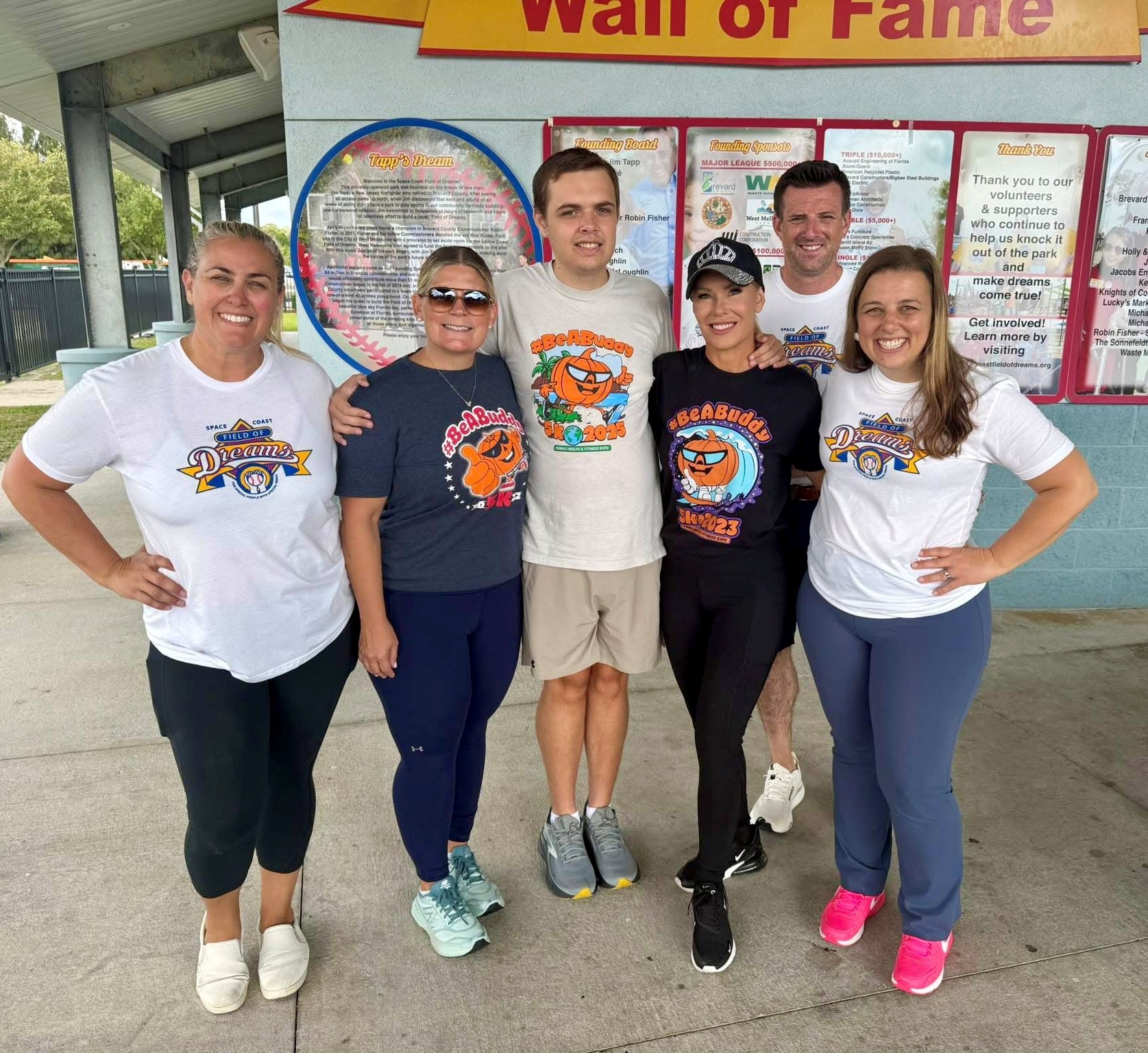 Six people standing together, smiling, in front of a wall with a display and text, some wearing matching shirts.