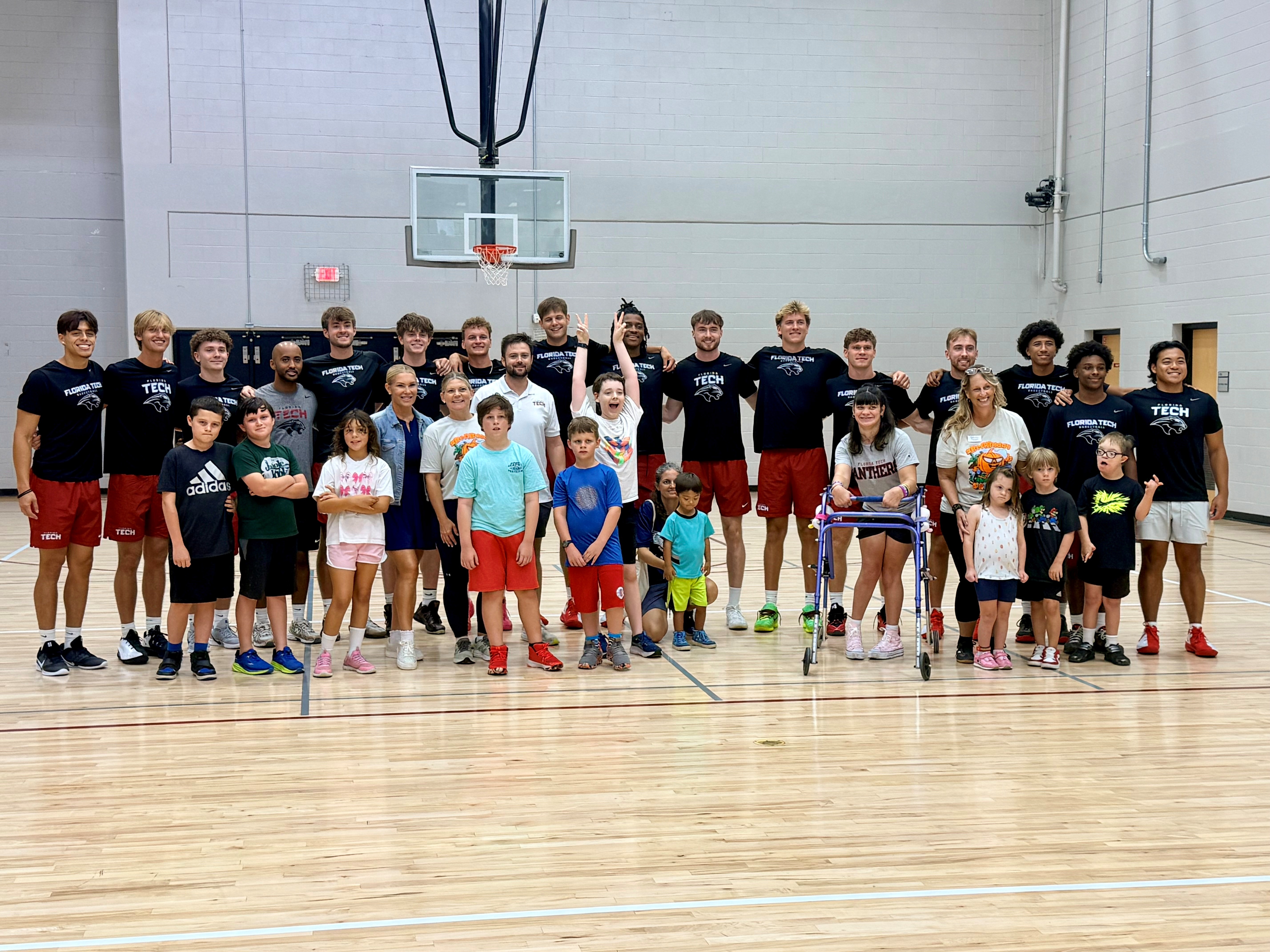 Group of children and adults standing on a basketball court, some holding basketballs, in a gymnasium.