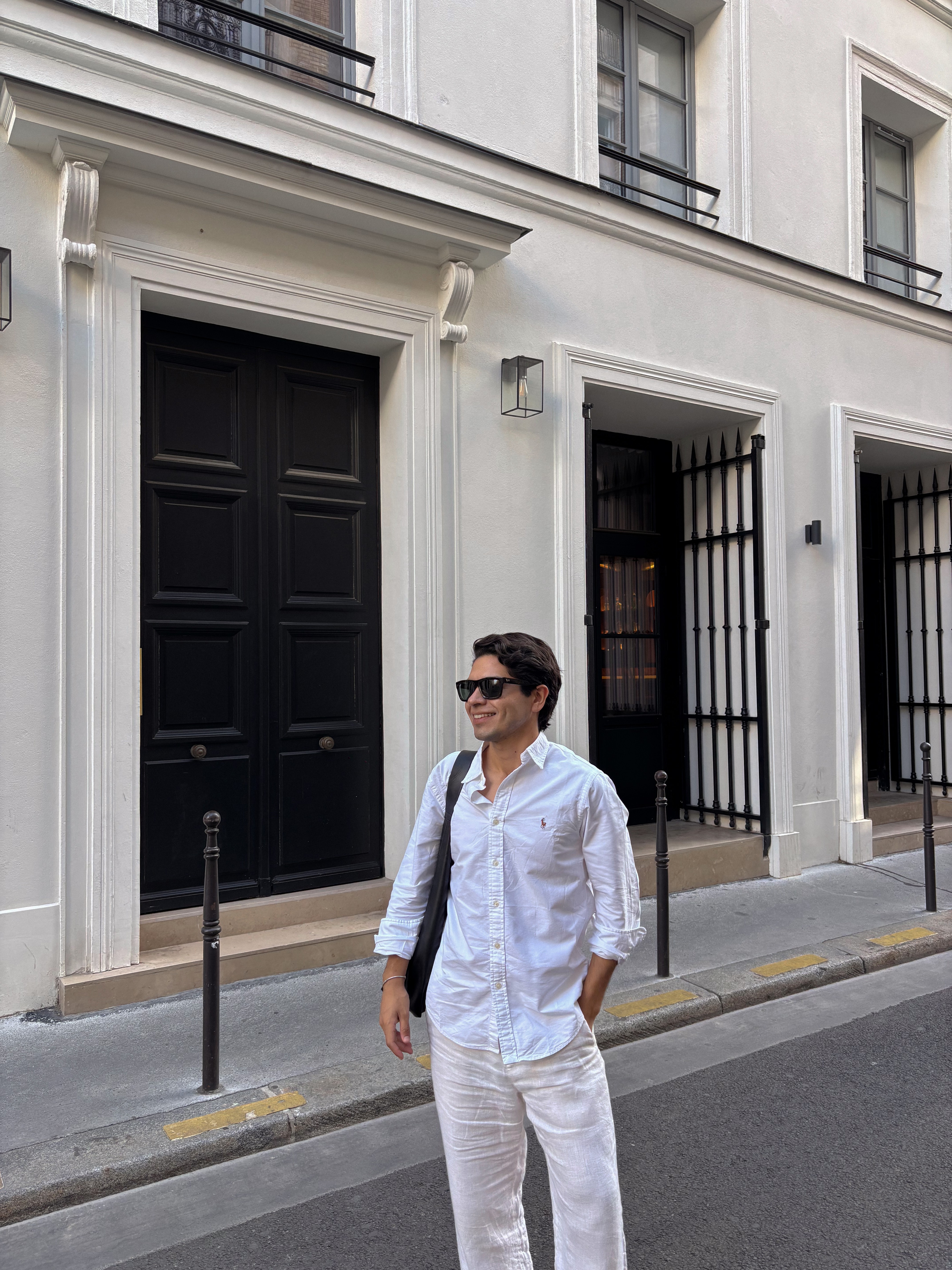 Person wearing sunglasses and white outfit standing on a city street in front of a white building with black doors.