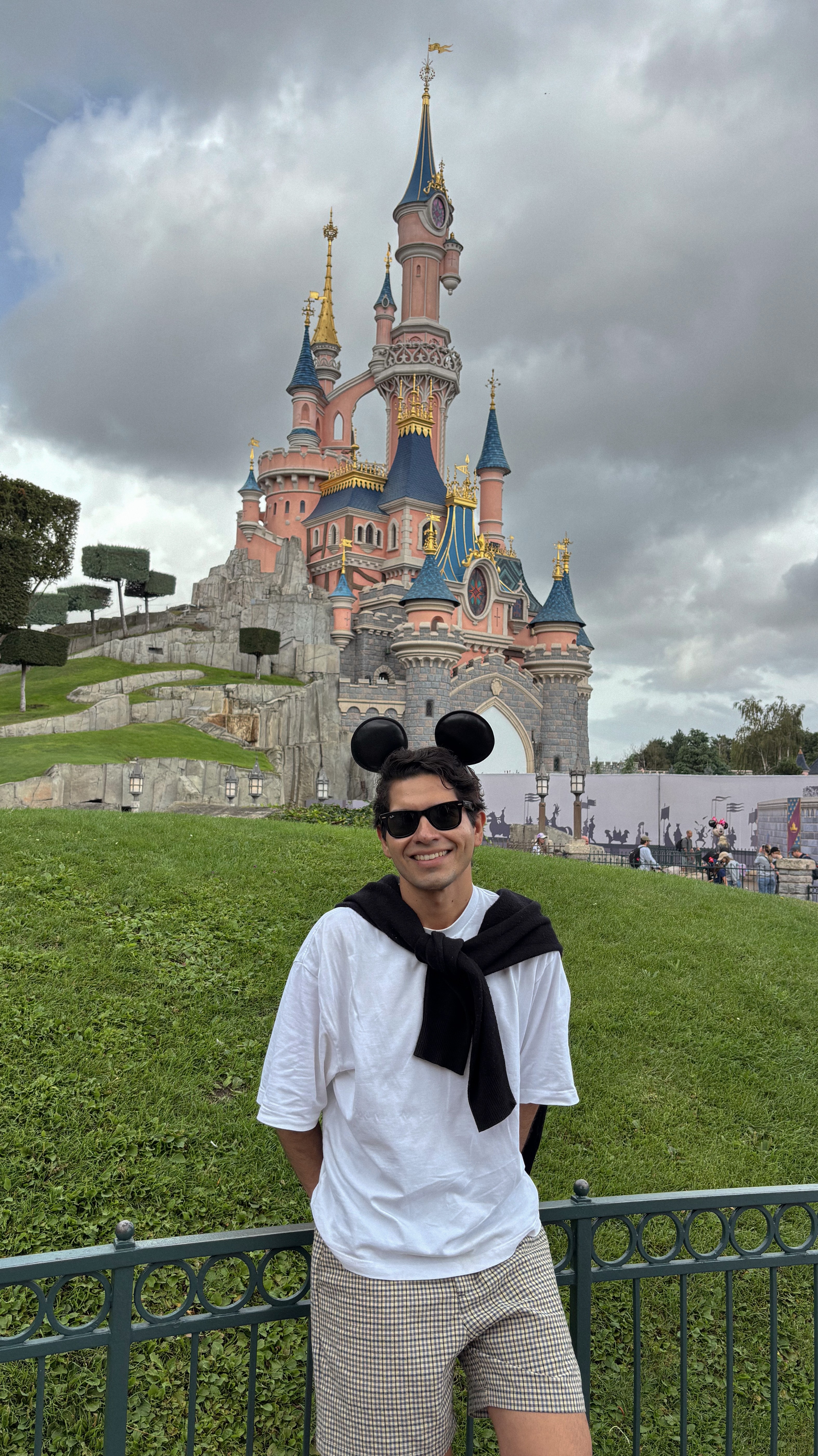 Young man wearing sunglasses, white shirt, and shorts standing in front of a castle with pink walls and blue spires.