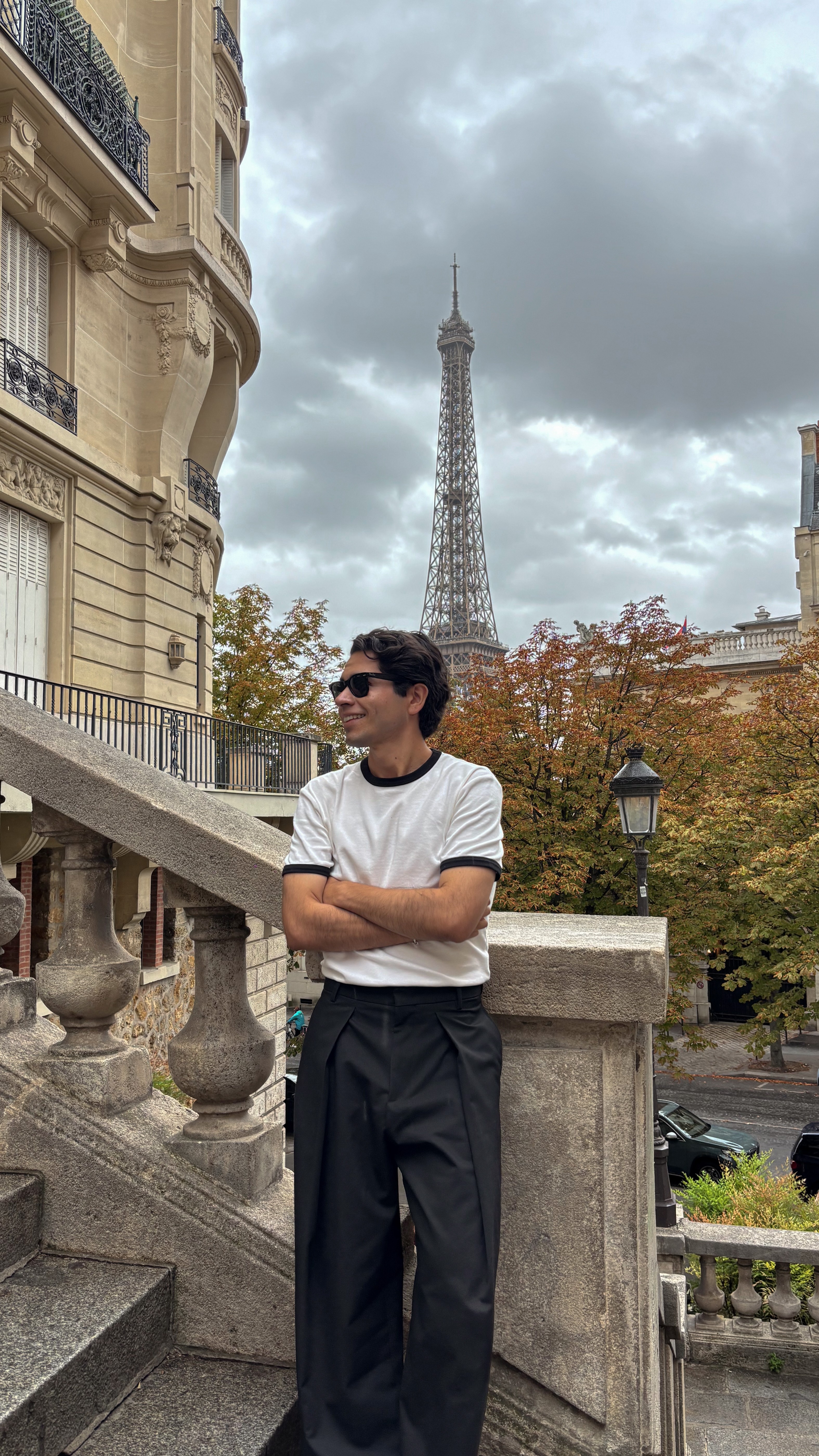 Young man with sunglasses standing outdoors near a stone railing, with the Eiffel Tower and cloudy sky in the background.