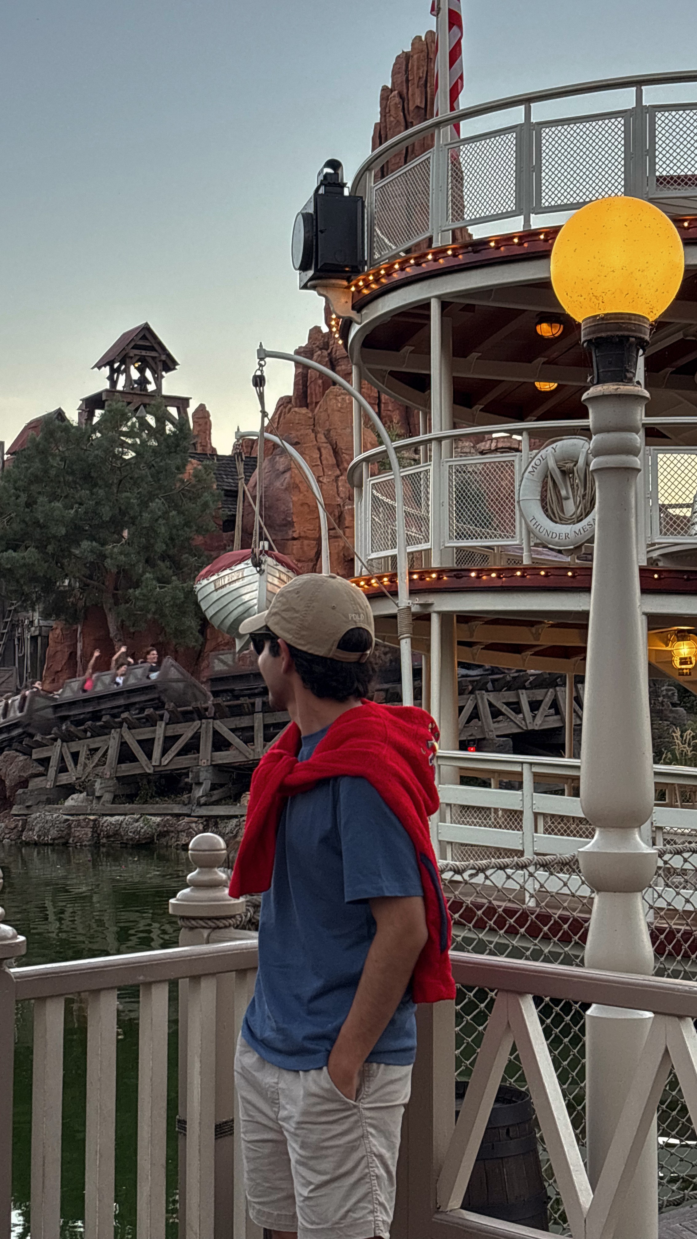 Person wearing a cap, red scarf, and blue shirt standing near a railing at an amusement park, with a building and water in background.