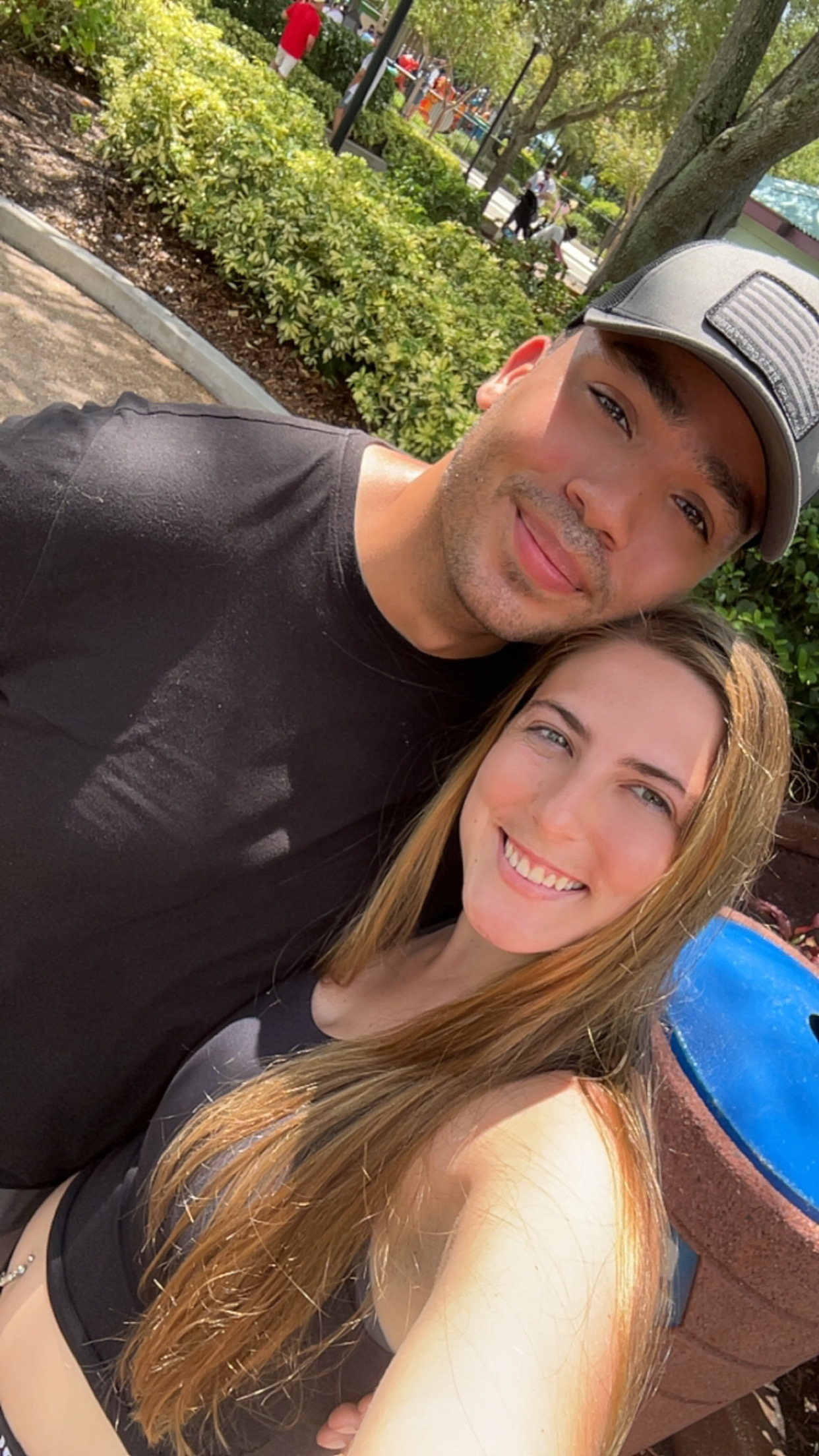 A smiling young man and woman taking a selfie outdoors with trees and a blue object in the background.