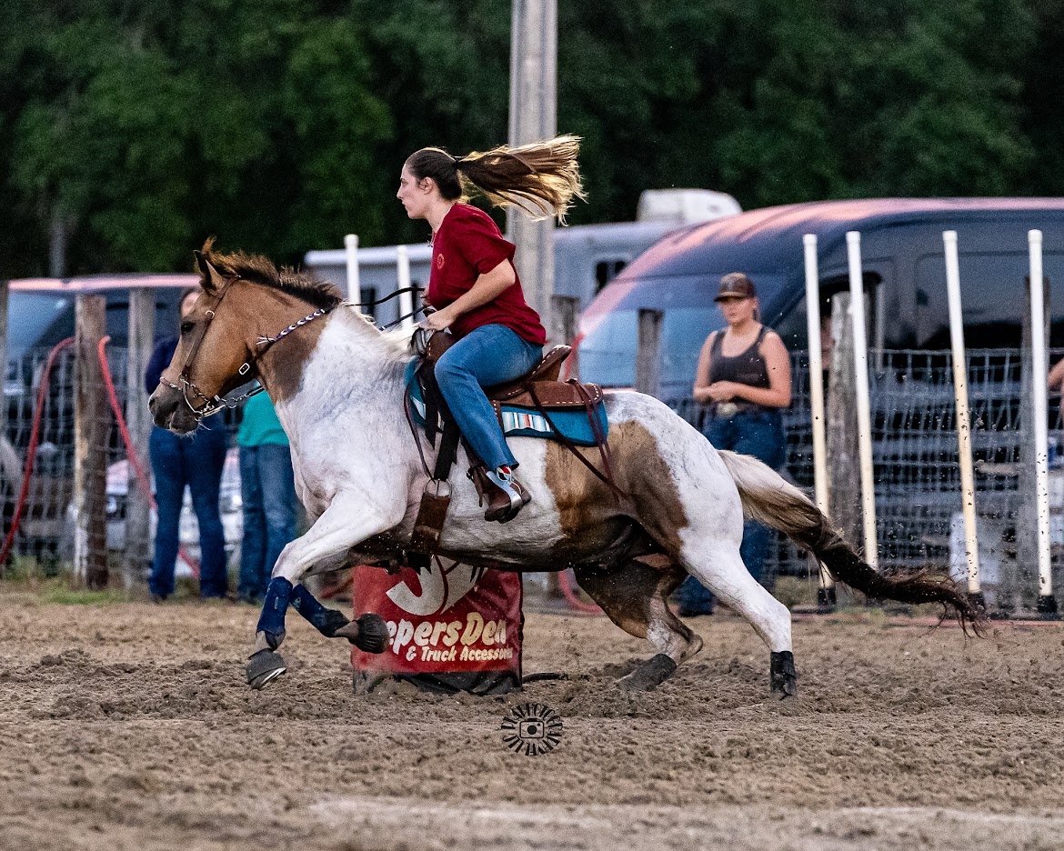 Young woman riding a galloping horse during a rodeo event, with spectators and vehicles in the background.
