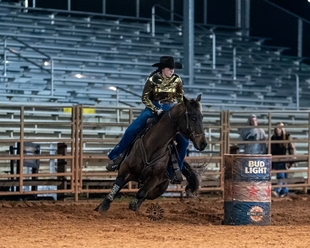 Person riding a horse around a barrel in an indoor arena, with empty bleachers in the background.