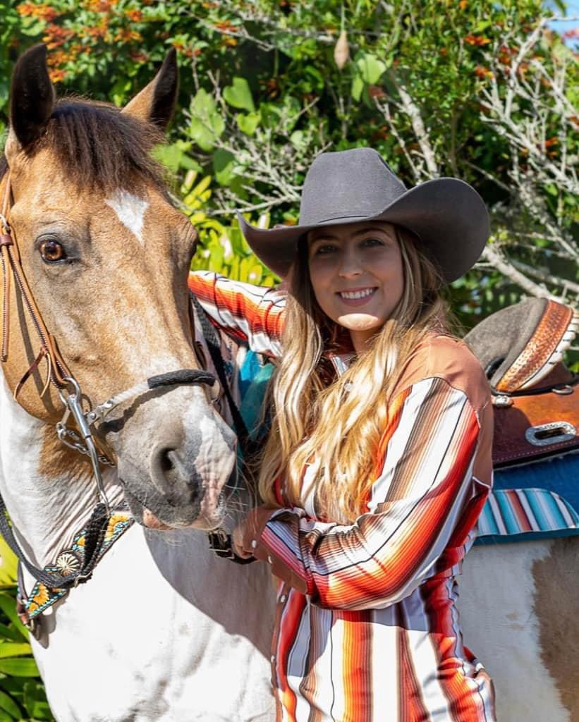 Young woman in a striped shirt and wide-brimmed hat smiling next to a horse outdoors with greenery.