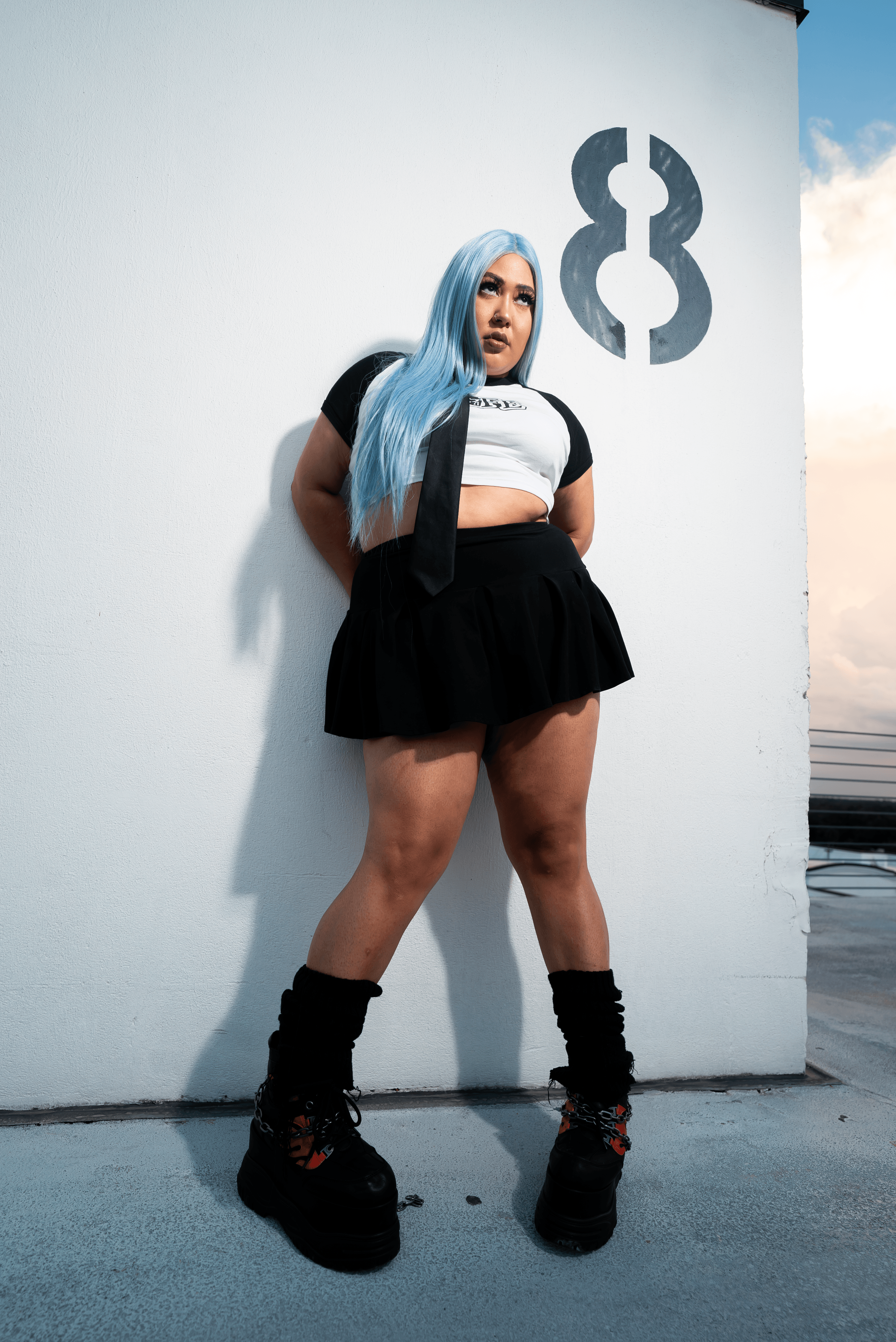 Young woman with long blue hair standing against a white wall with a large number 8, wearing a black skirt and sneakers.