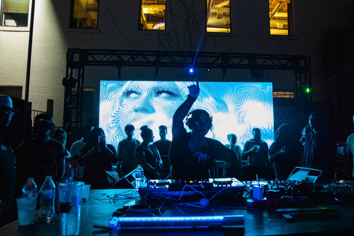 Crowd of people at a nighttime outdoor event with a DJ and a large screen displaying a face, some holding drinks.