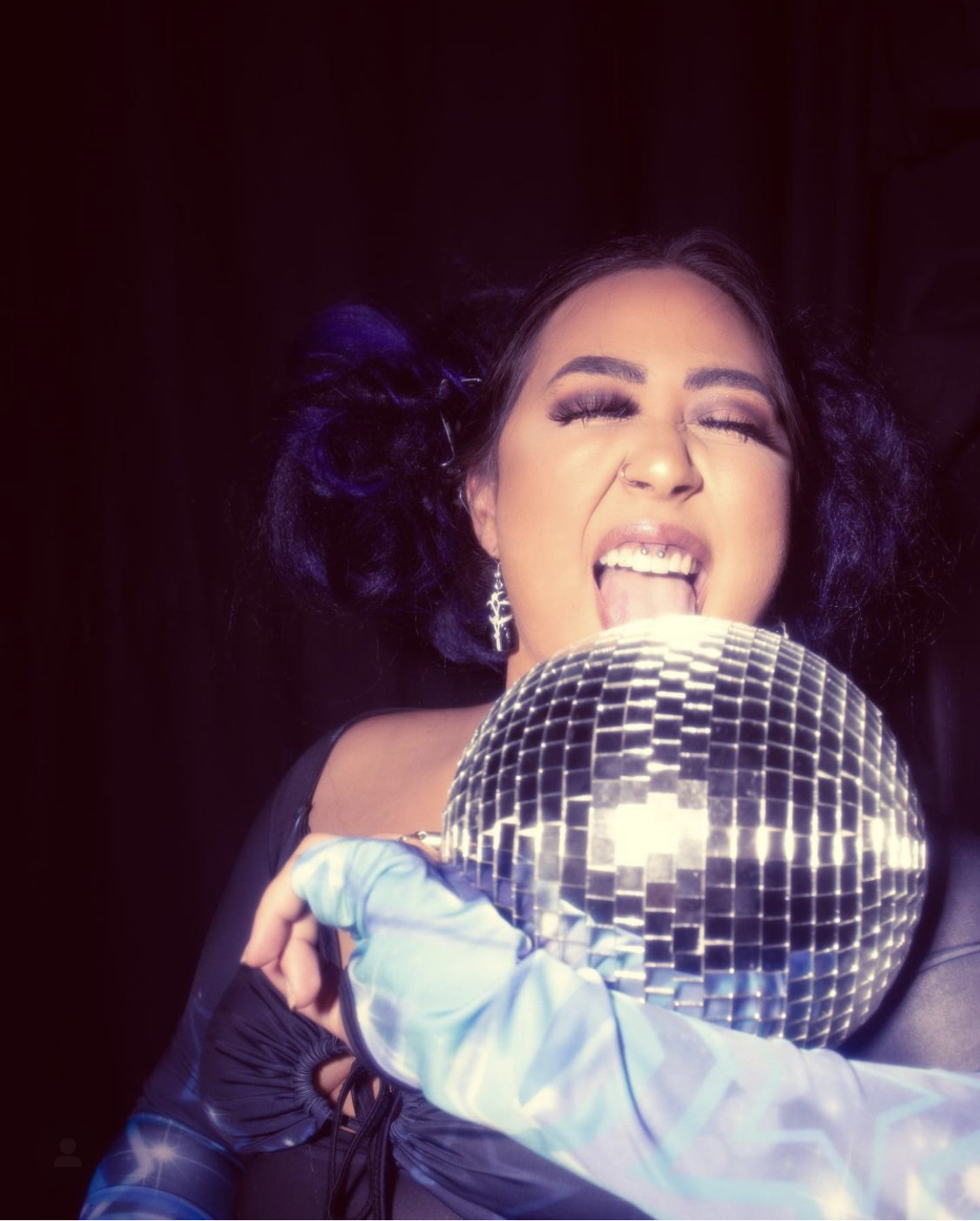 Person with dark curly hair holding a disco ball, smiling with eyes closed, dark background, wearing earrings and a strapless top.