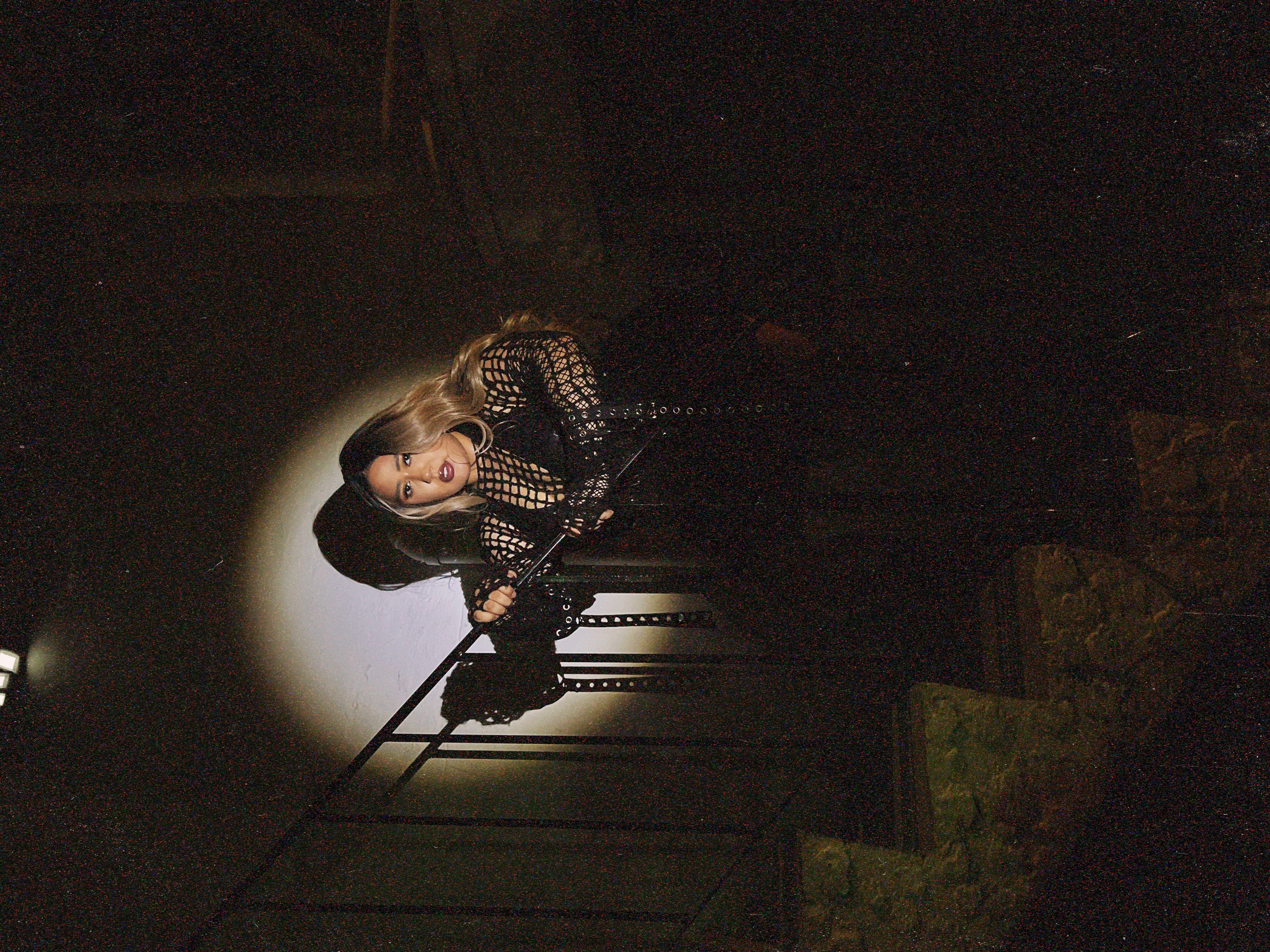 Person with long hair and dark clothing stands on stairs in a spotlight, holding a microphone, in a dark setting.