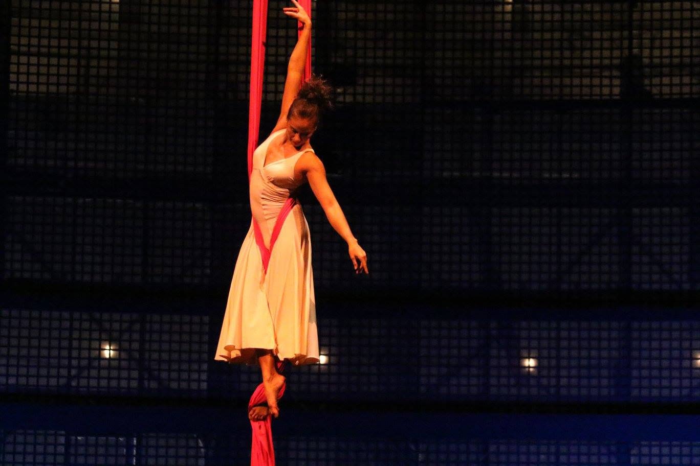 A performer hangs from a red fabric in a dark stage setting, wearing a light-colored dress with a red sash.