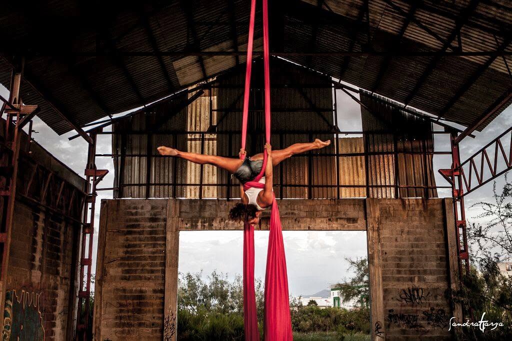 Person performing aerial silk pose upside down in a barn-like structure with open sides and trees outside.